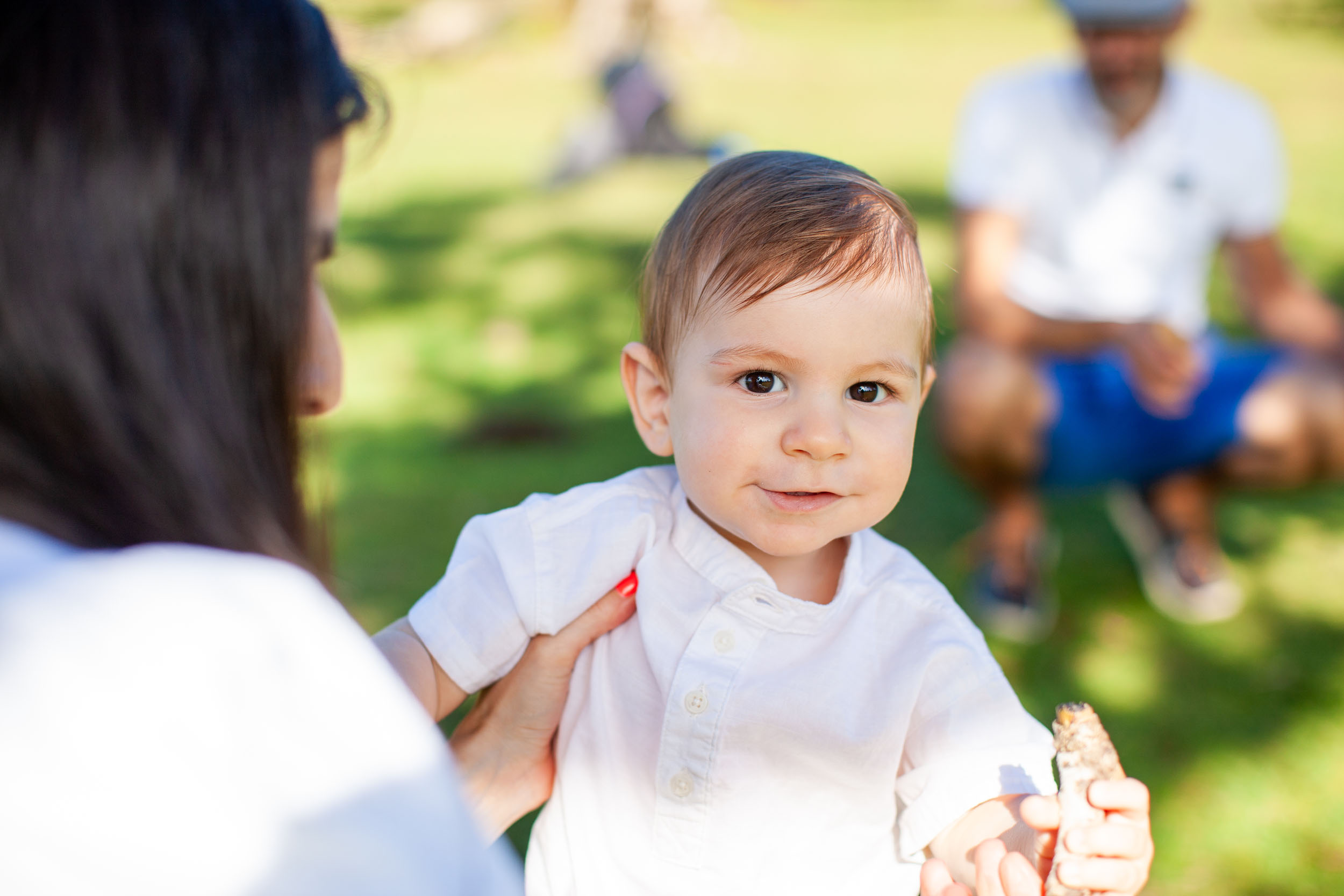 Close portrait of baby boy looking directly at camera, mother's hand on his shoulder, father softly blurred in background, Fanal Forest, Madeira