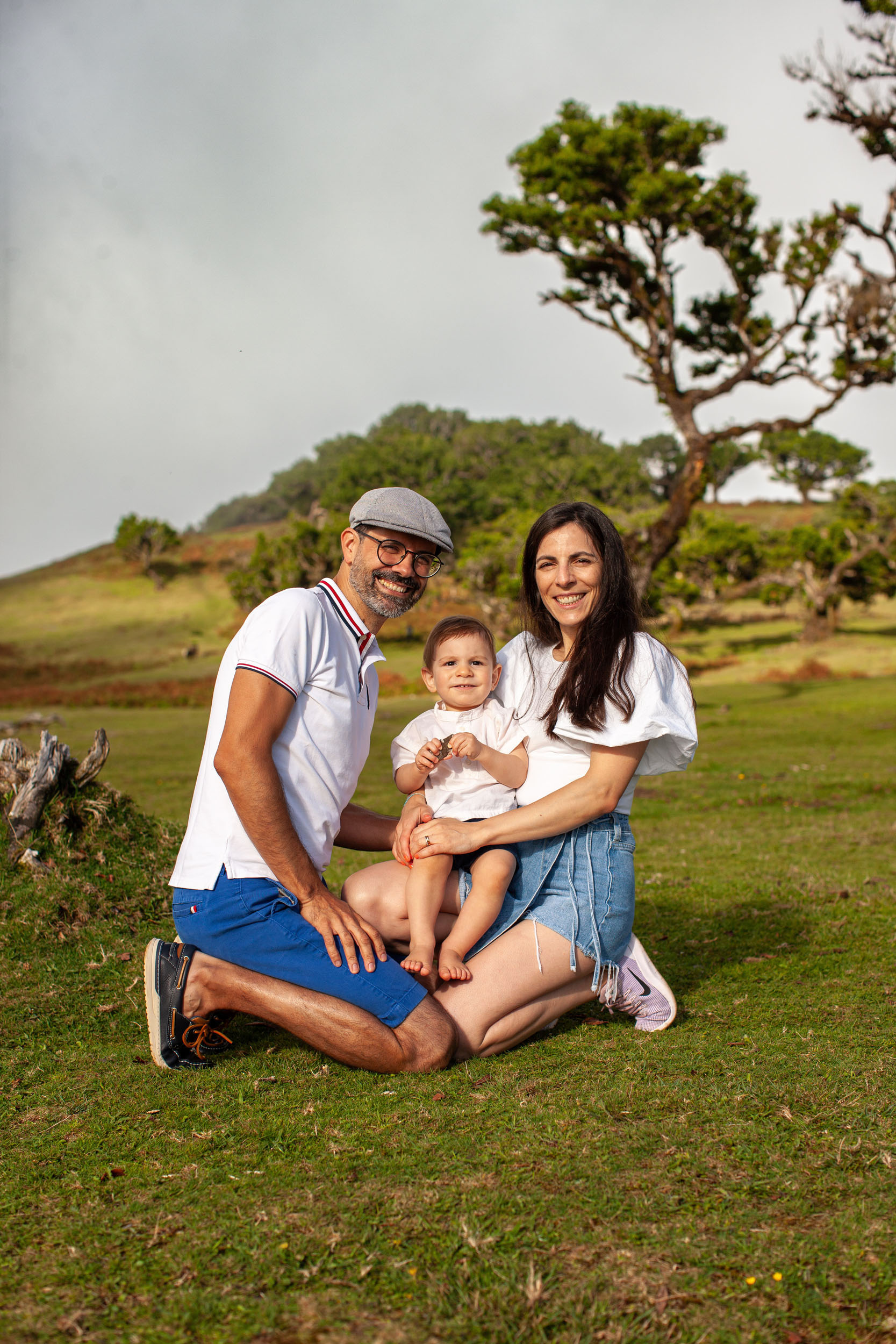 Family seated on the grass, baby on mother's lap, father kneeling beside, ancient laurel tree on hillside behind, Fanal Forest, Madeira