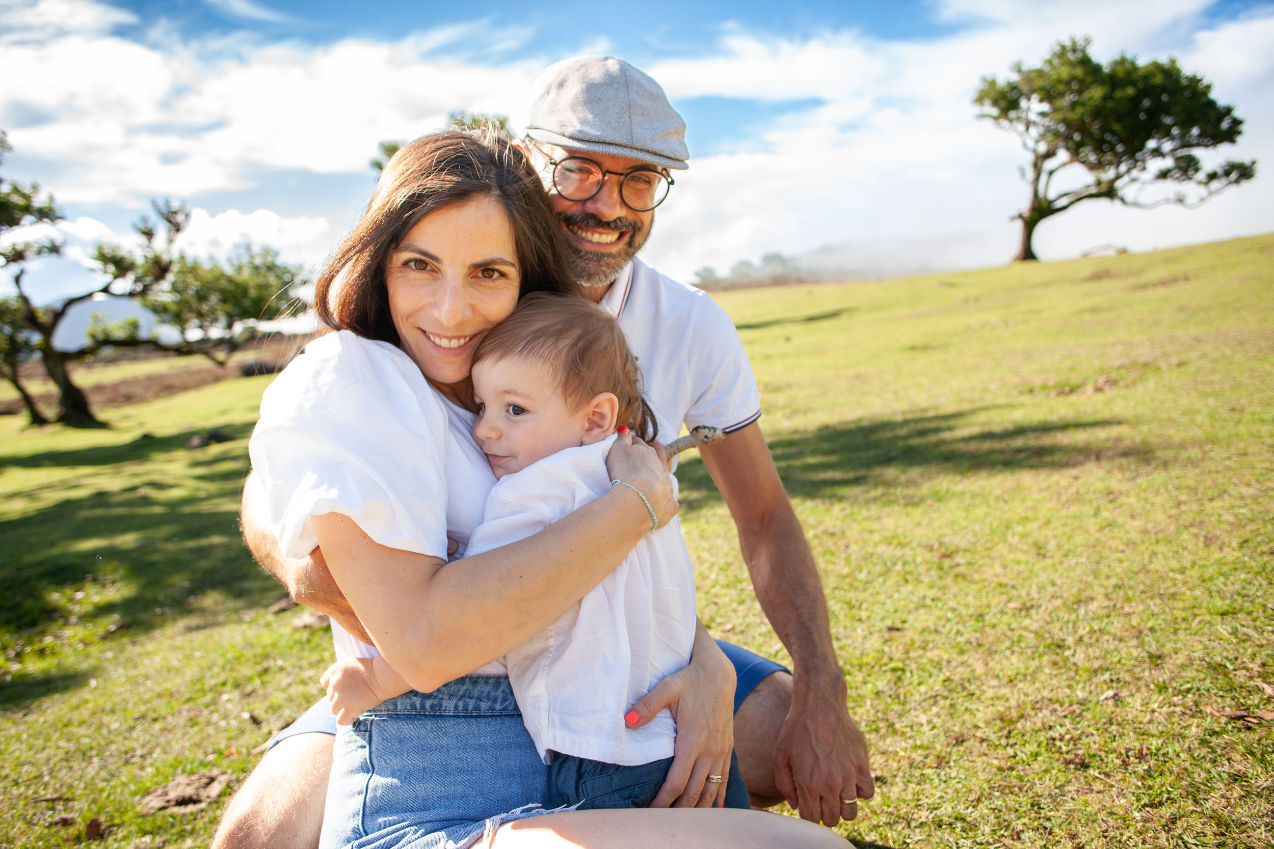 Family portrait on the grass, mother hugging baby, father alongside, laurel trees and clouds behind, Fanal Forest, Madeira