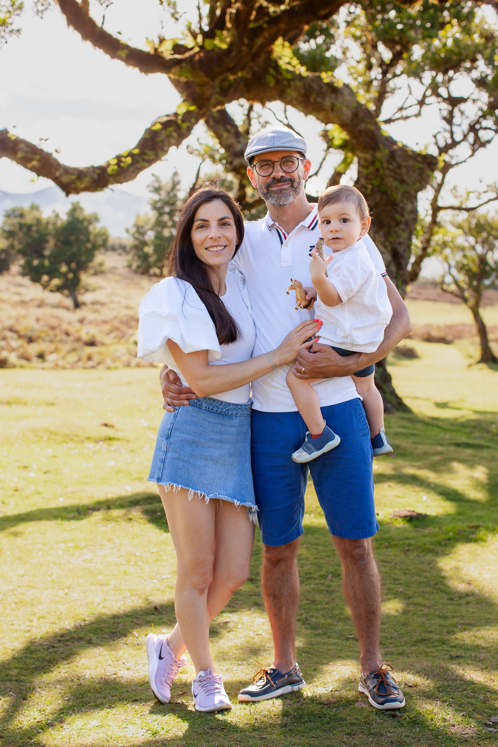 Family portrait standing in warm light, father holding baby with toy horse, mother beside him, laurel tree behind, Fanal Forest, Madeira