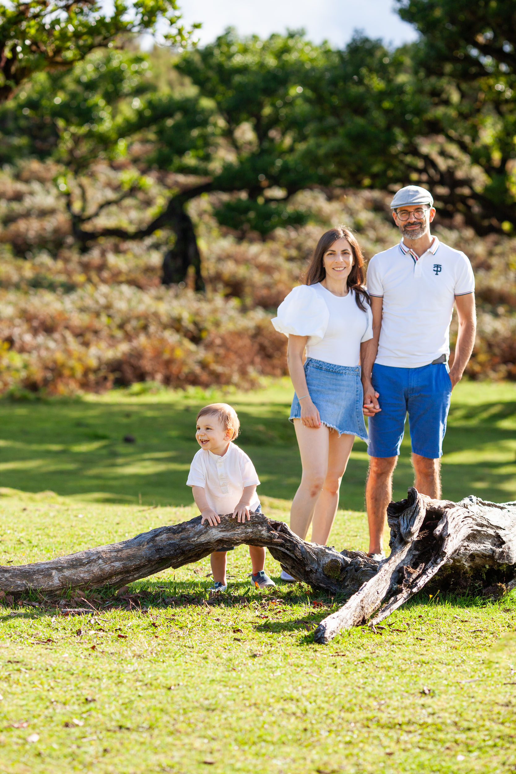 Family of three by a fallen laurel log, baby climbing on it, parents standing behind, forest in warm light, Fanal Forest, Madeira
