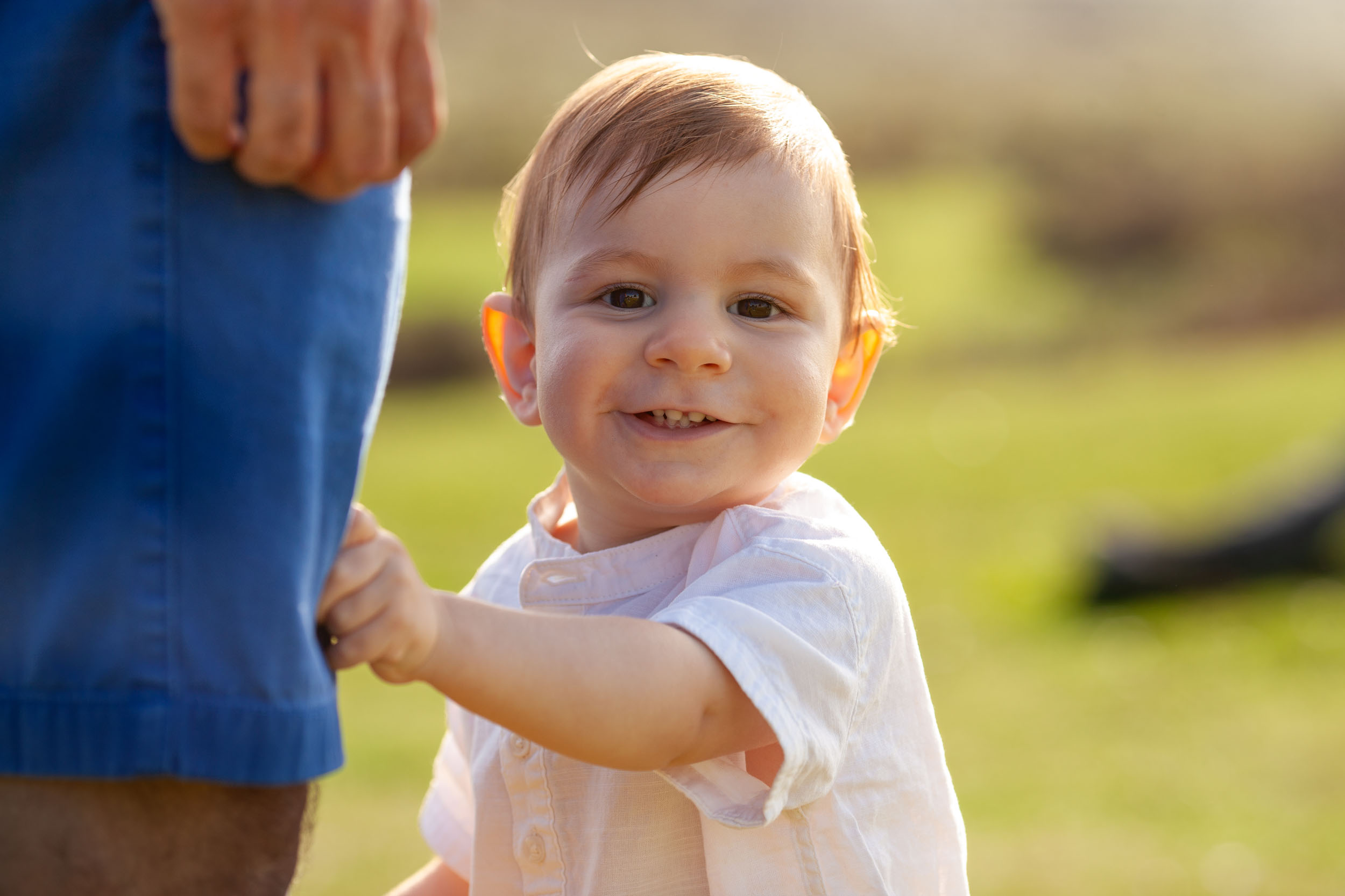 Close portrait of baby boy smiling broadly at camera, holding father's shorts, golden backlight, Fanal meadow behind, Madeira