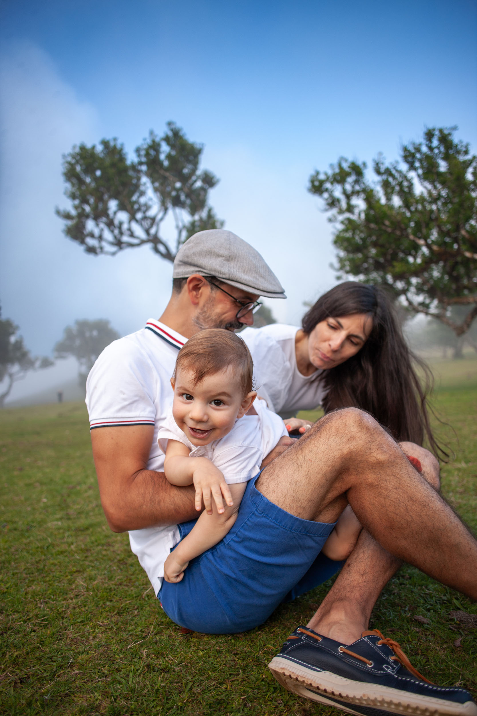 Family sitting on grass, father hugging baby, mother leaning in gently, fog and laurel trees behind, Fanal Forest, Madeira