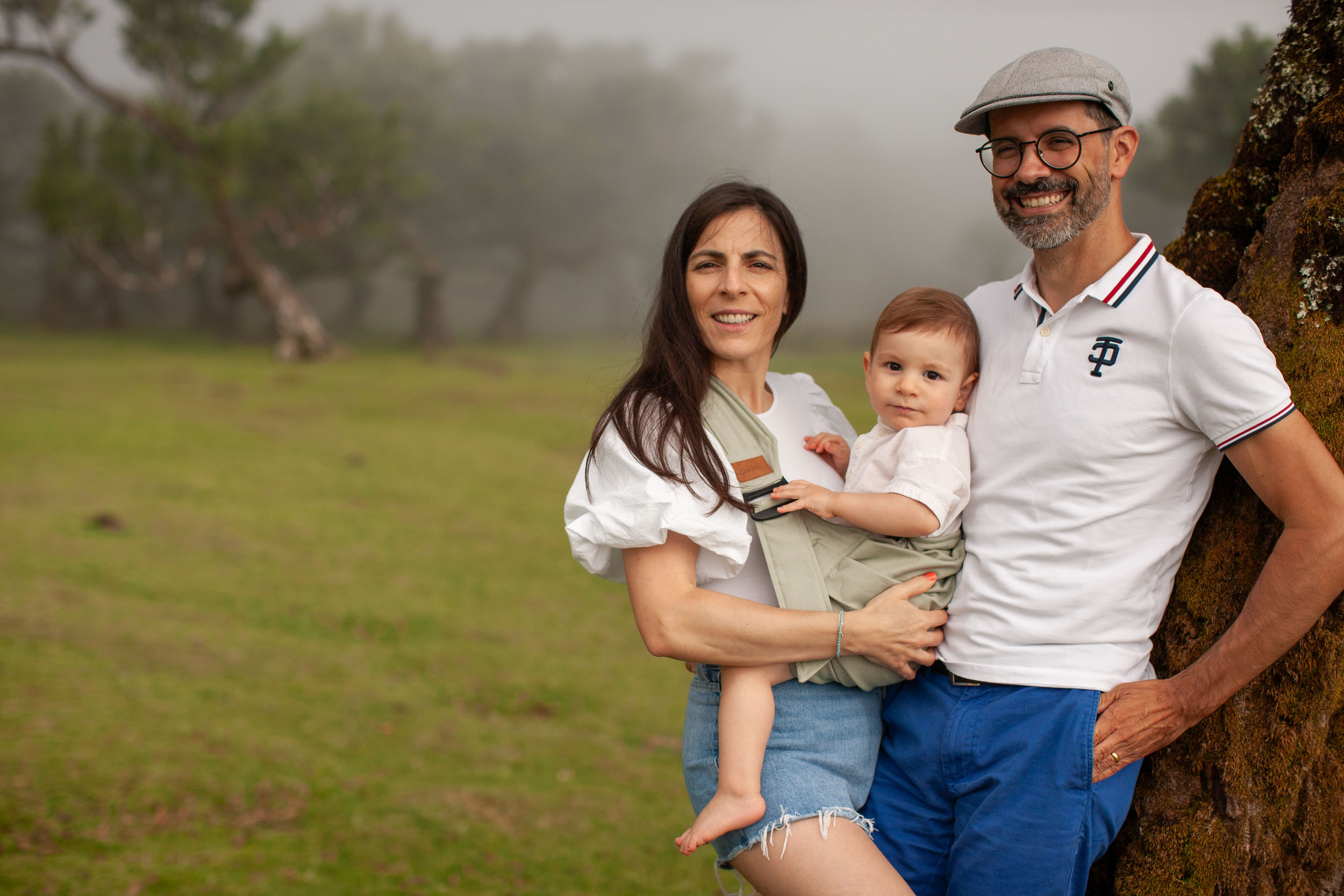 Family portrait leaning against an ancient laurel tree, foggy green meadow behind, mother holding baby, both parents smiling, Fanal Forest, Madeira