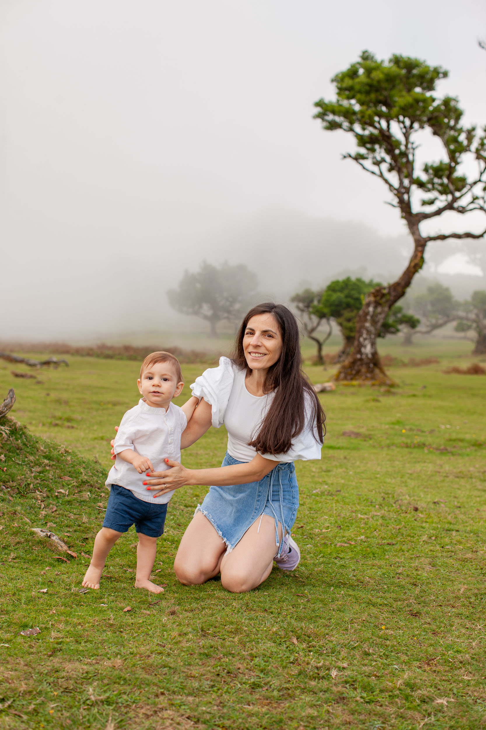 Mother kneeling beside standing barefoot baby on green grass, foggy ancient laurel trees behind, Fanal Forest, Madeira