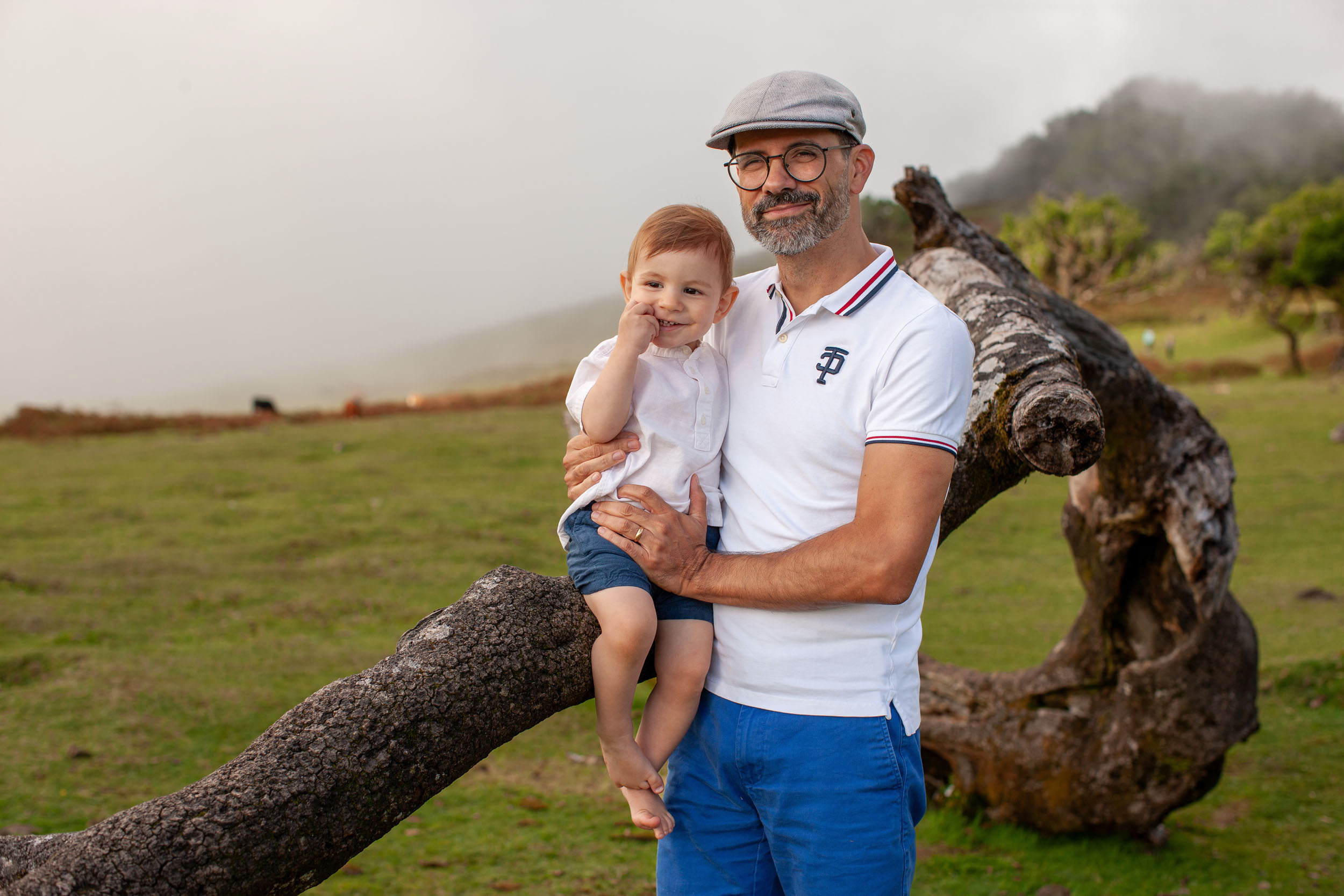 Father holding baby boy on his arm beside a fallen laurel branch, foggy plateau and mountains behind, Fanal Forest, Madeira