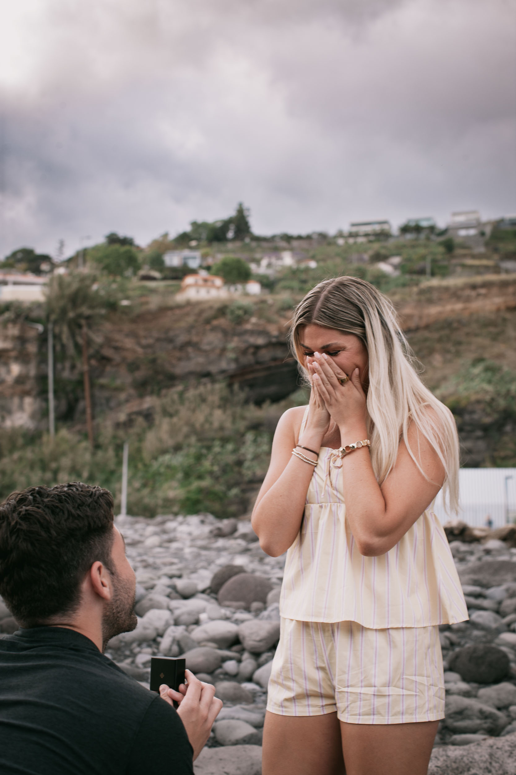 Amelie covering her mouth and nose with both hands at the moment of the proposal, Mike's kneeling shoulder in the foreground left, overcast sky over the green Funchal cliff at Praia Formosa, Madeira