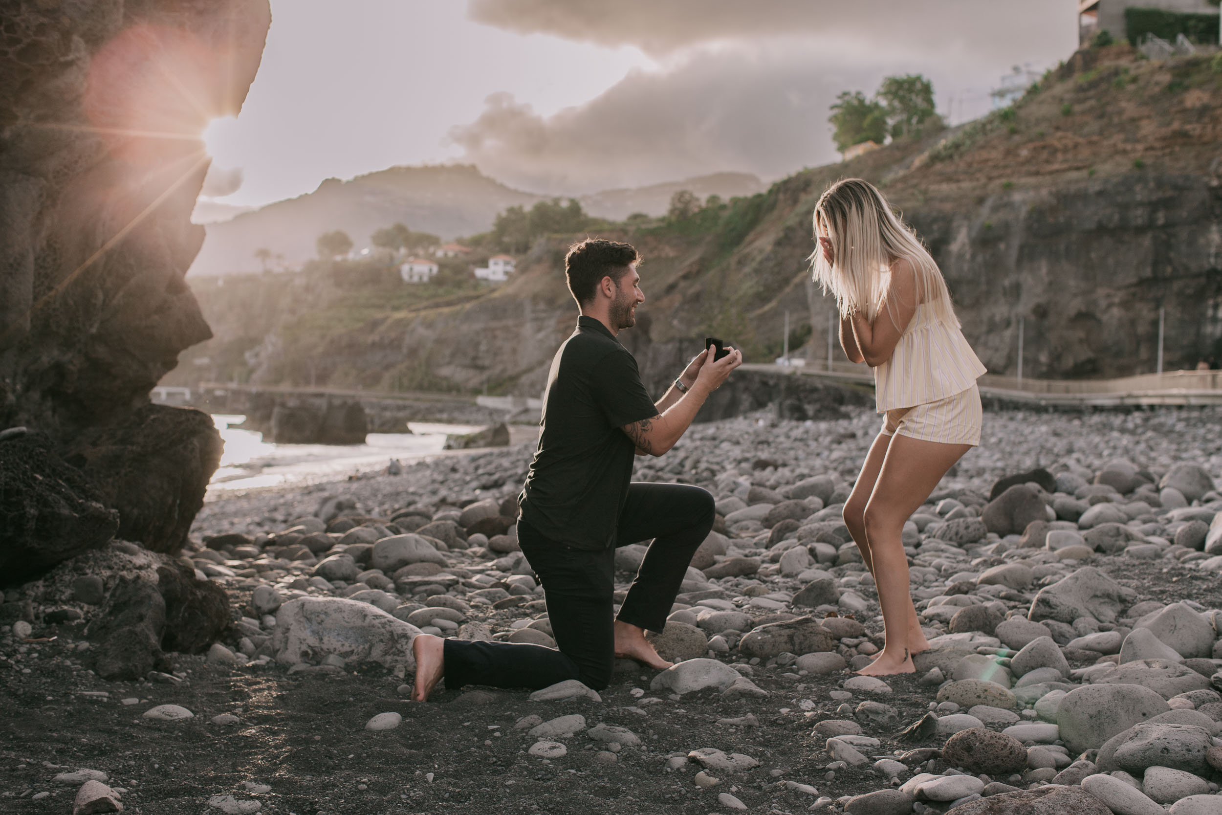 Mike still kneeling on one knee offering the open ring box, Amelie standing barefoot with her hair falling forward, sun flare beside the dark rock face at Praia Formosa, Madeira