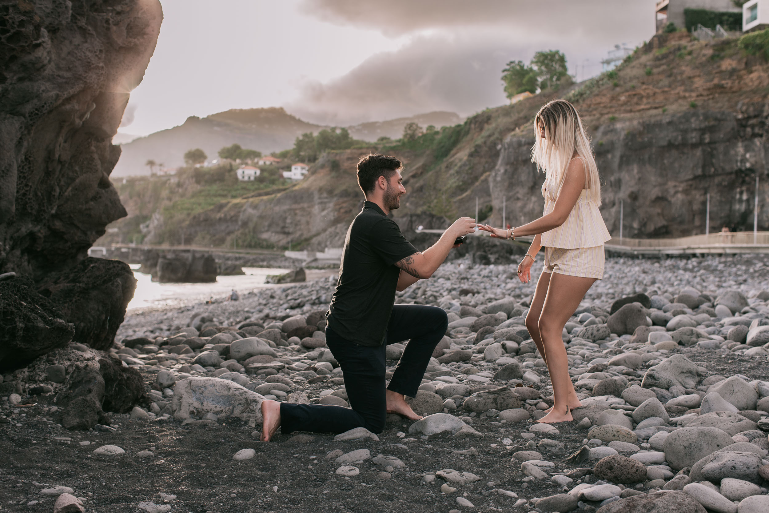 Mike sliding the engagement ring onto Amelie's outstretched hand from one knee on the boulder beach, large sea stack to the left at Praia Formosa, Madeira