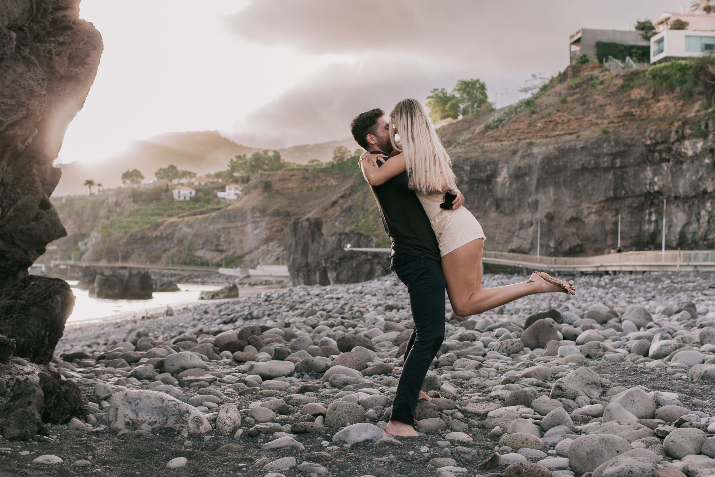 Mike lifting Amelie clean off her feet in the first kiss after she said yes, her foot kicked back in the air and her arms around his neck, sea stack on the left at Praia Formosa, Madeira