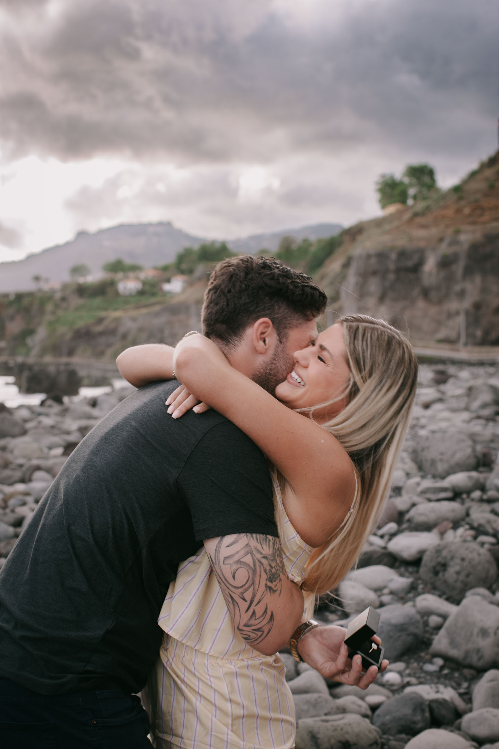 Mike pressing his face into Amelie's chest in a tight hug, her arms around his head as she smiles with eyes closed, his hand holding the open ring box behind her back, dark storm clouds and his tribal sleeve tattoo, Praia Formosa, Madeira