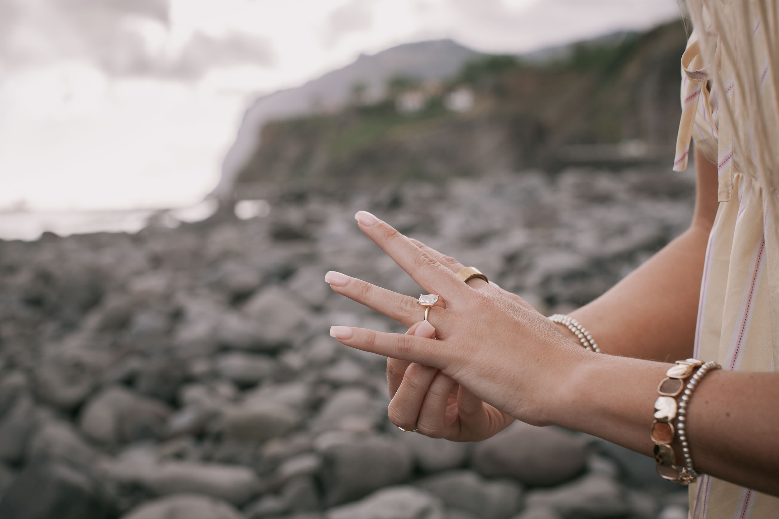 Tight detail of Amelie's new emerald-cut solitaire engagement ring on her finger, her hand held up against Mike's hand with her stacked gold-and-stone bracelets visible, blurred boulder shore behind at Praia Formosa, Madeira