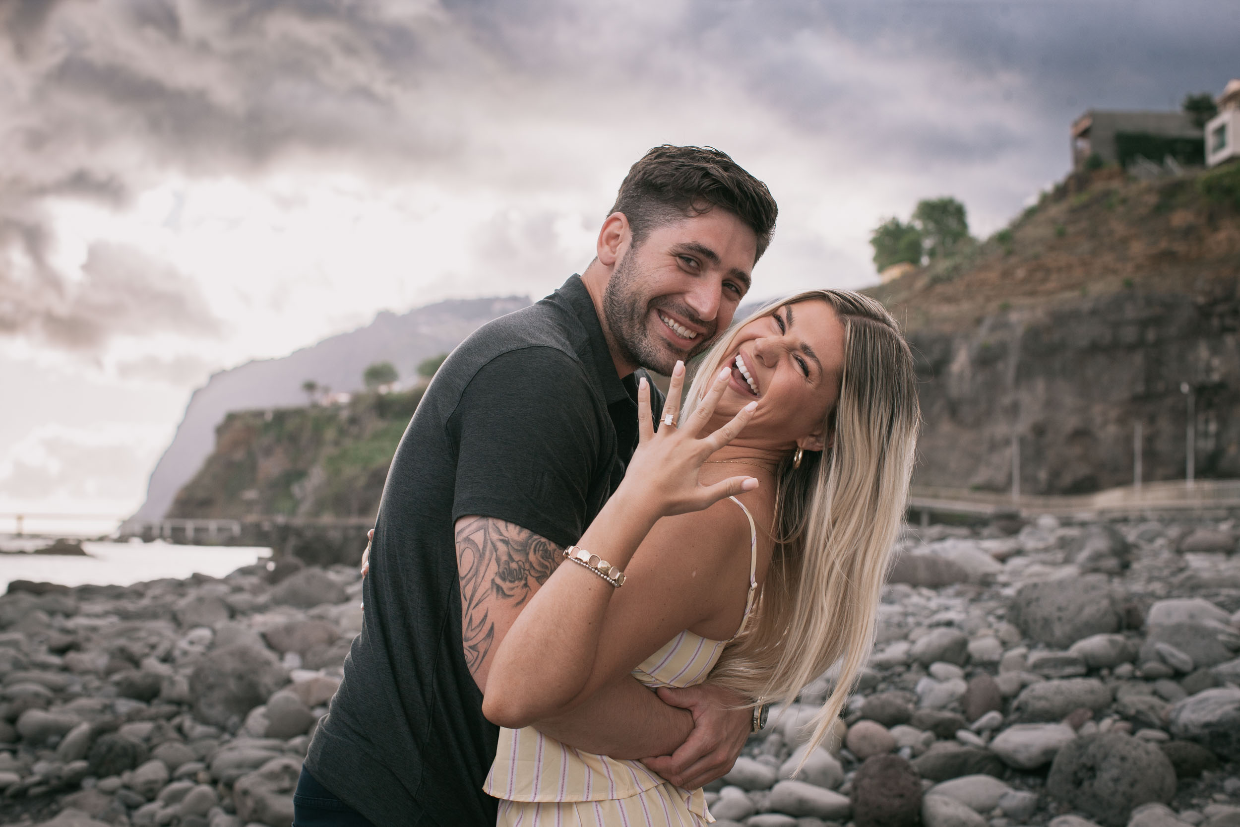 Mike holding Amelie close from the side as both turn to laugh into the camera, her ringed hand held out high to show the new engagement ring, Cabo Girão cliff softened behind them at Praia Formosa, Madeira