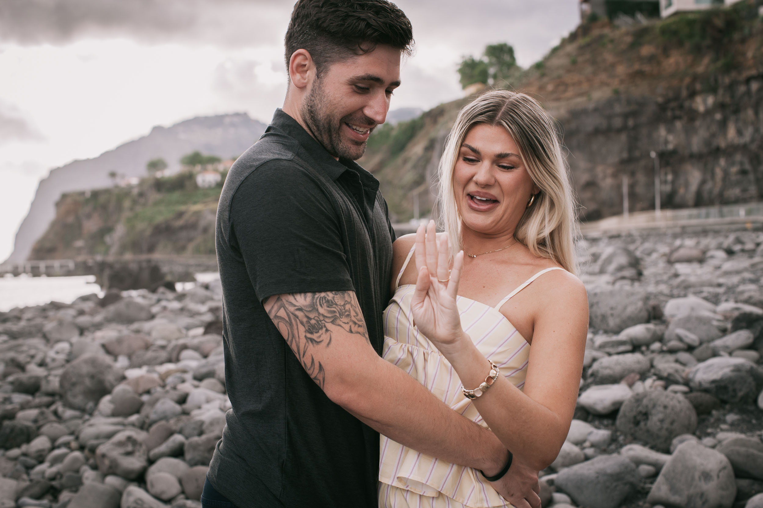 Mike looking down at Amelie's raised hand admiring the new ring as she scrunches her face in disbelief, both standing on the boulder beach with Cabo Girão behind, Praia Formosa, Madeira