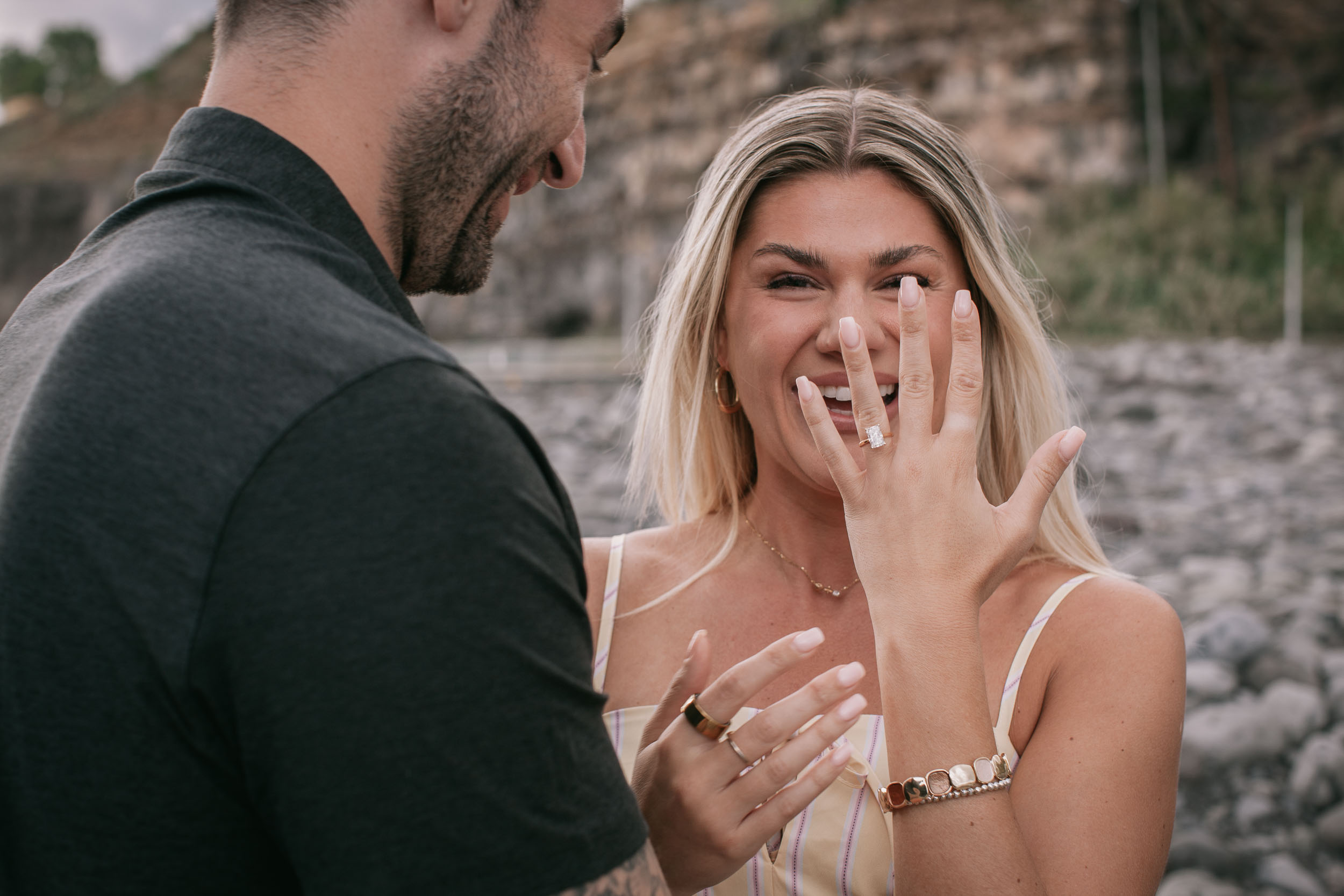 Amelie laughing with one hand to her cheek and the other held up showing the ring, Mike's smiling face in soft foreground from behind, Praia Formosa, Madeira