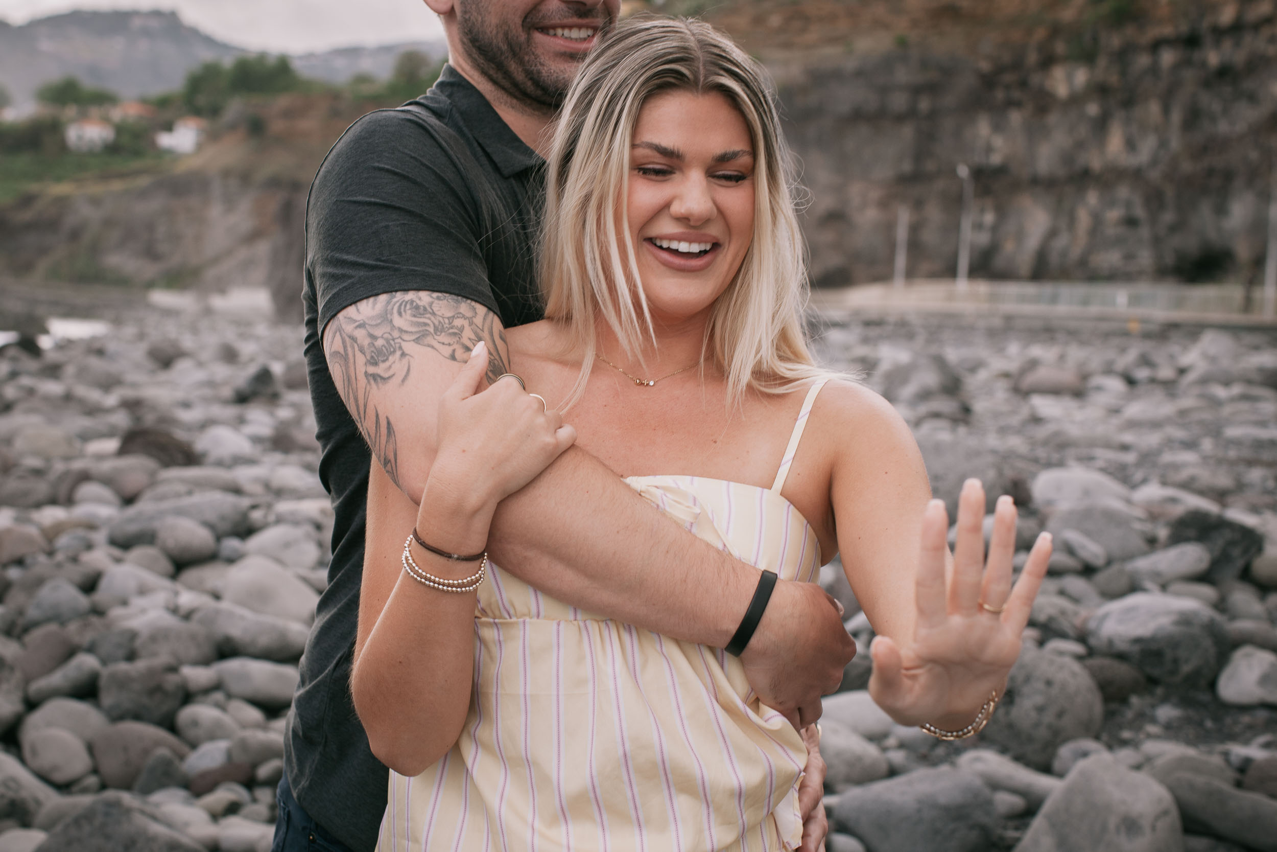 Mike wrapping his tattooed arms around Amelie from behind as she laughs and holds her ringed hand out to one side, her black watch and her ring catching the light, Praia Formosa, Madeira