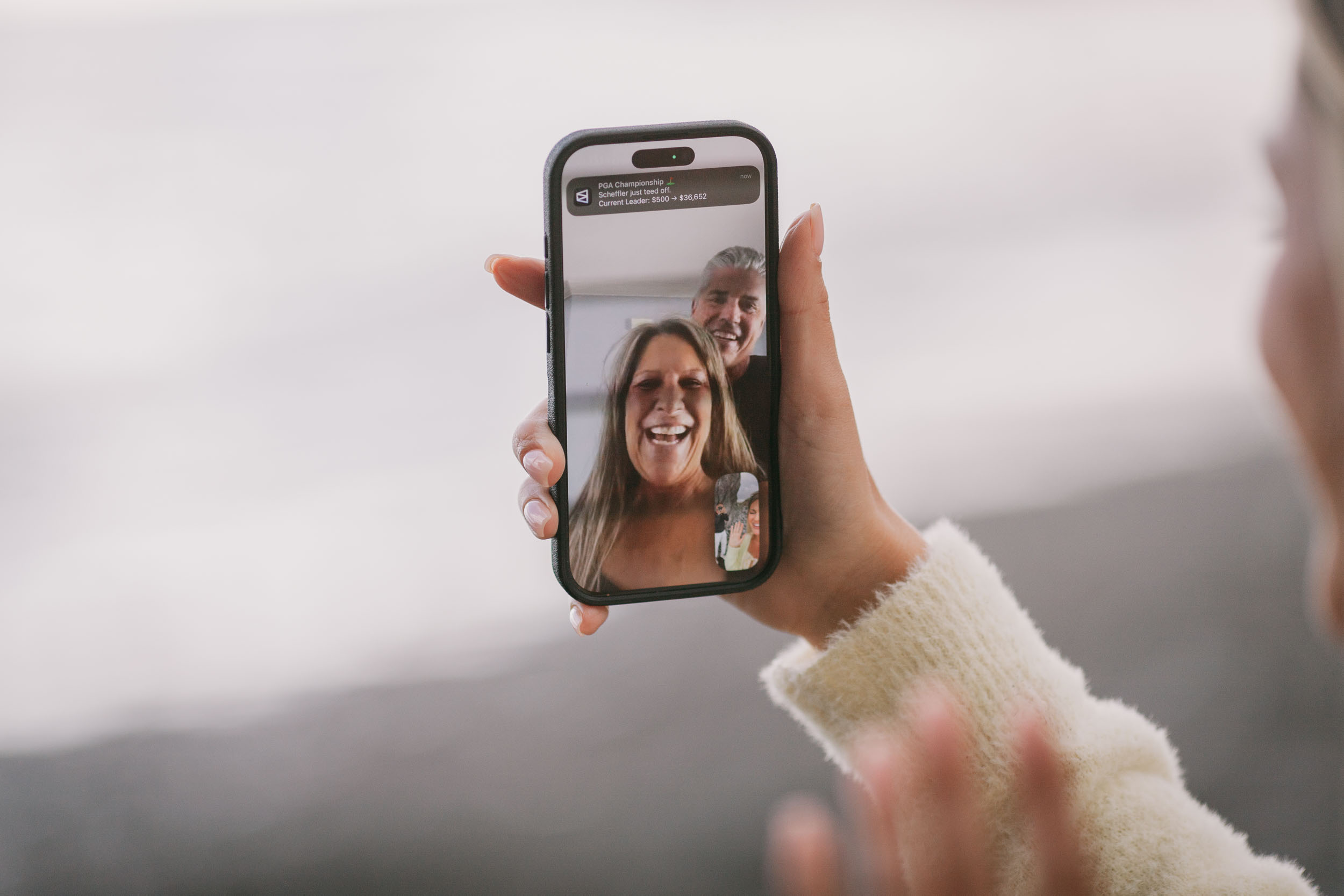 Amelie holding her phone up showing a video call with her grinning parents, the fresh engagement ring on her finger and a white fuzzy sweater on her sleeve, Praia Formosa, Madeira
