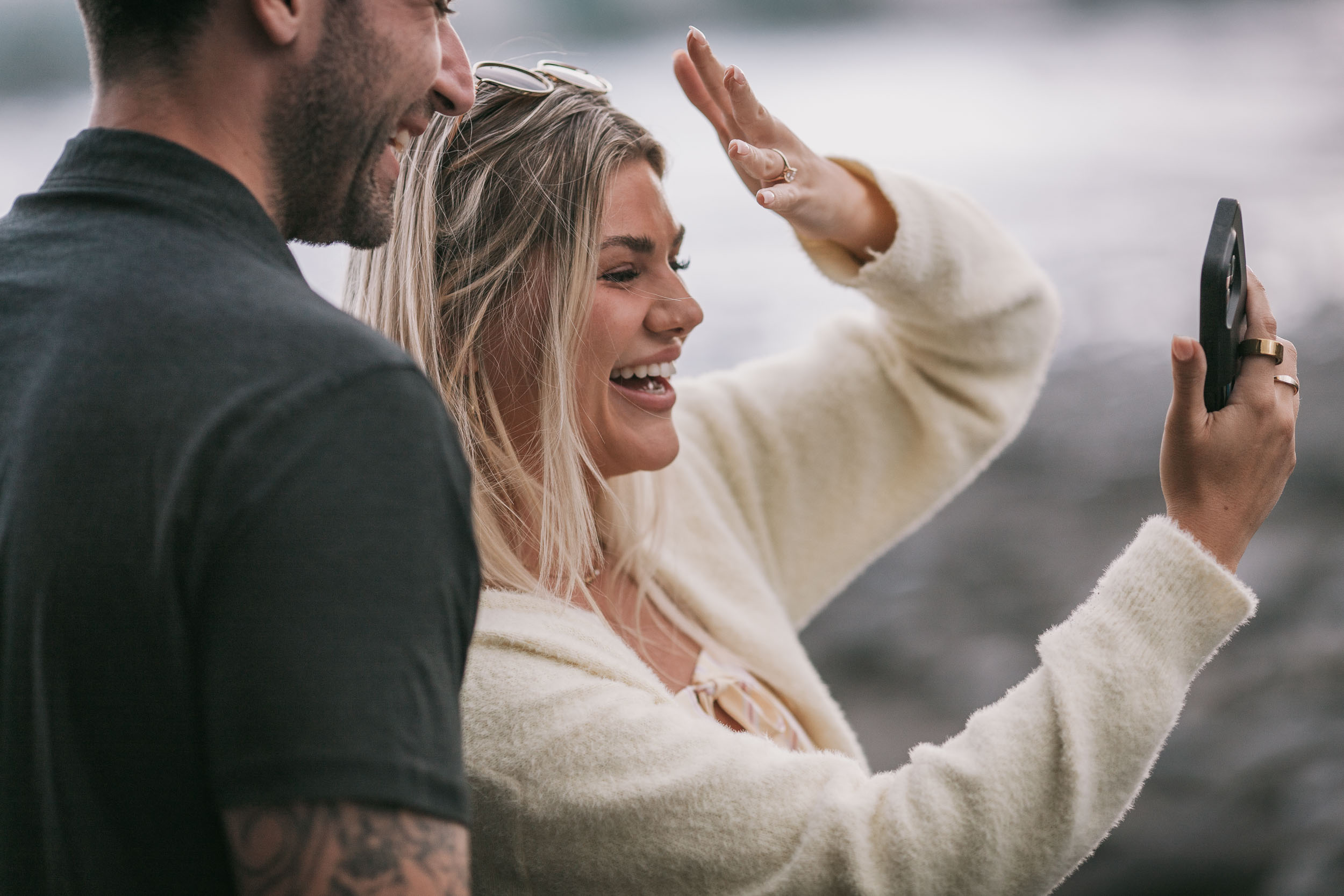 Side view of Amelie waving toward the phone she holds out at arm's length, Mike grinning beside her with sunglasses pushed up on her head, both seen in profile at Praia Formosa, Madeira