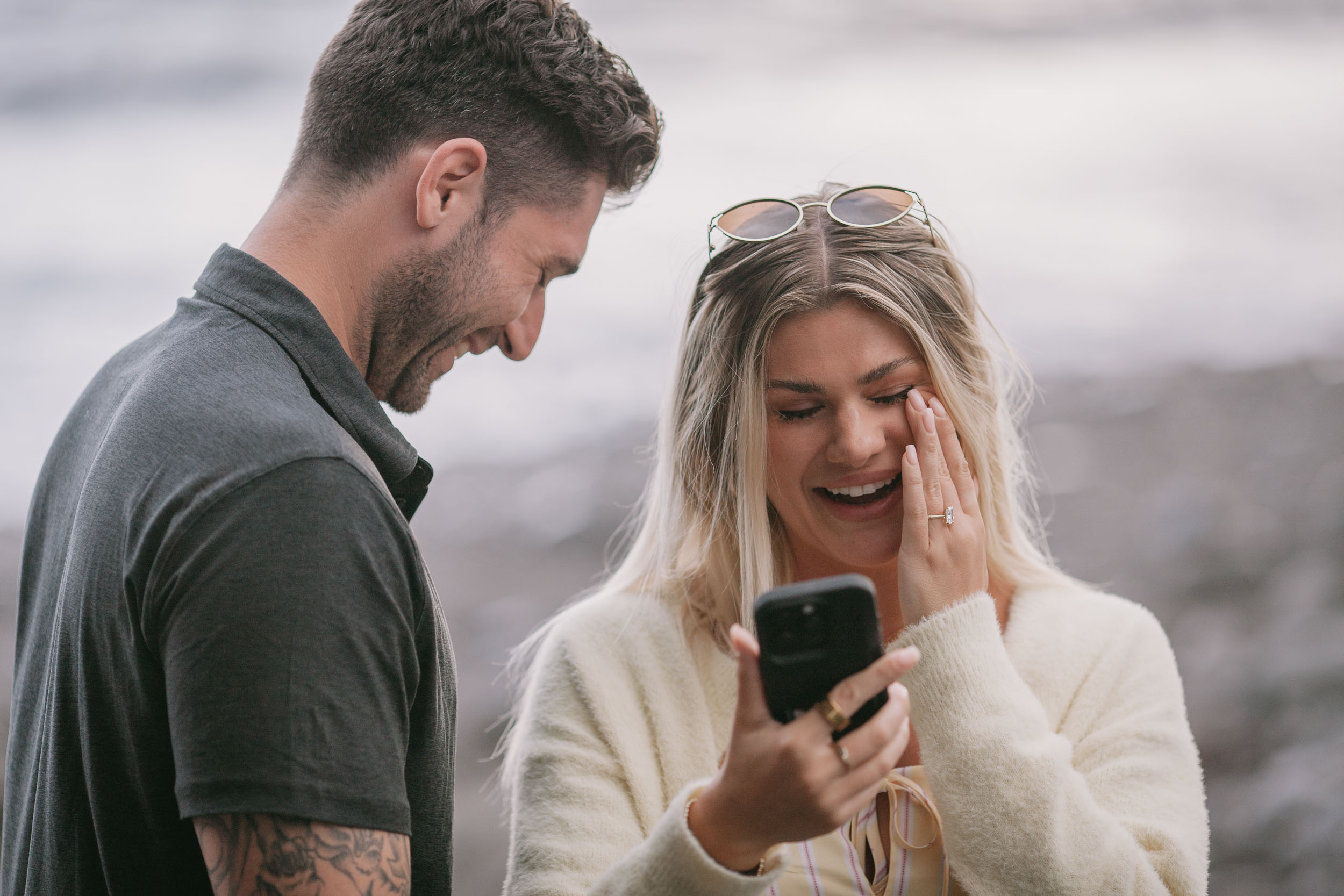 Amelie wiping a tear under her eye while looking down at the phone, Mike laughing in profile beside her, sunglasses on top of her head and the new ring visible on her hand, Praia Formosa, Madeira