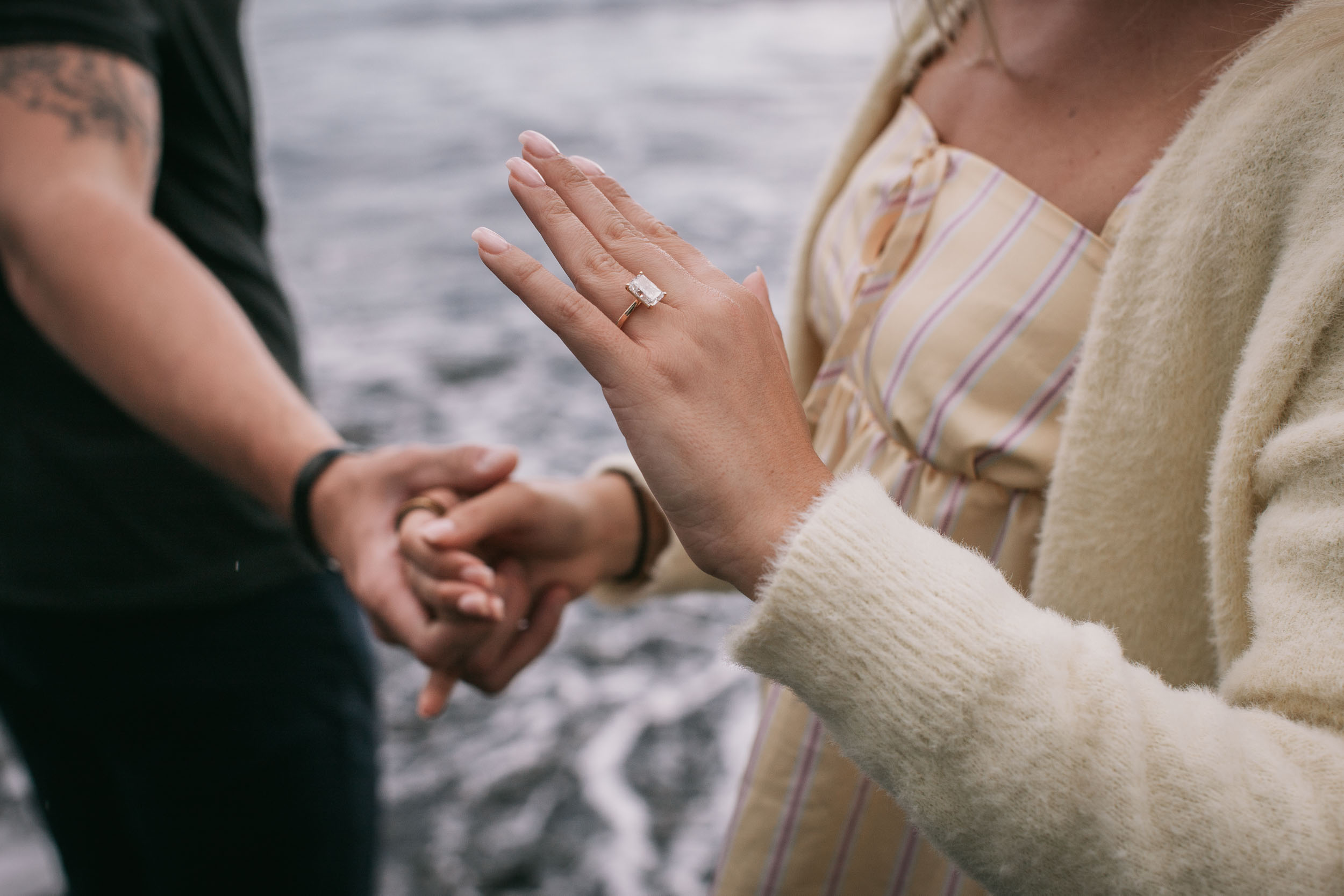 Close detail of Amelie's hand resting flat over Mike's clasped hands, her ring catching the daylight against the blurred grey surf, Praia Formosa, Madeira