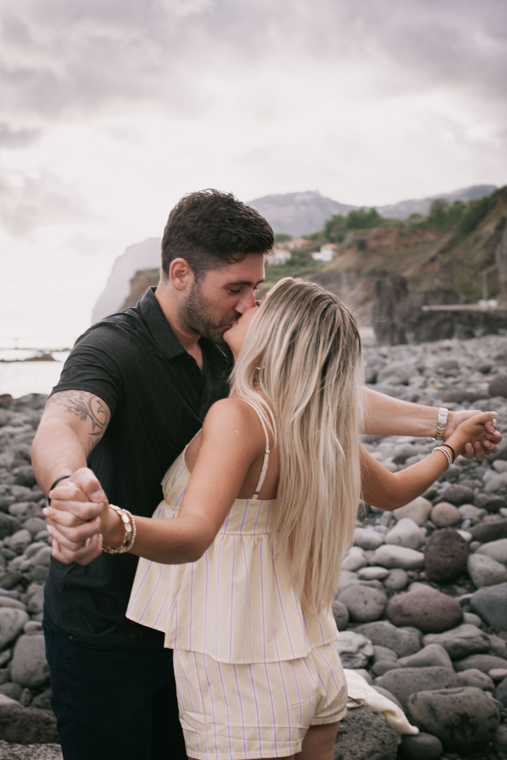 Mike and Amelie kissing standing up on the wet black sand, mountains and the long Funchal coastal bridge stretching across the background at Praia Formosa, Madeira