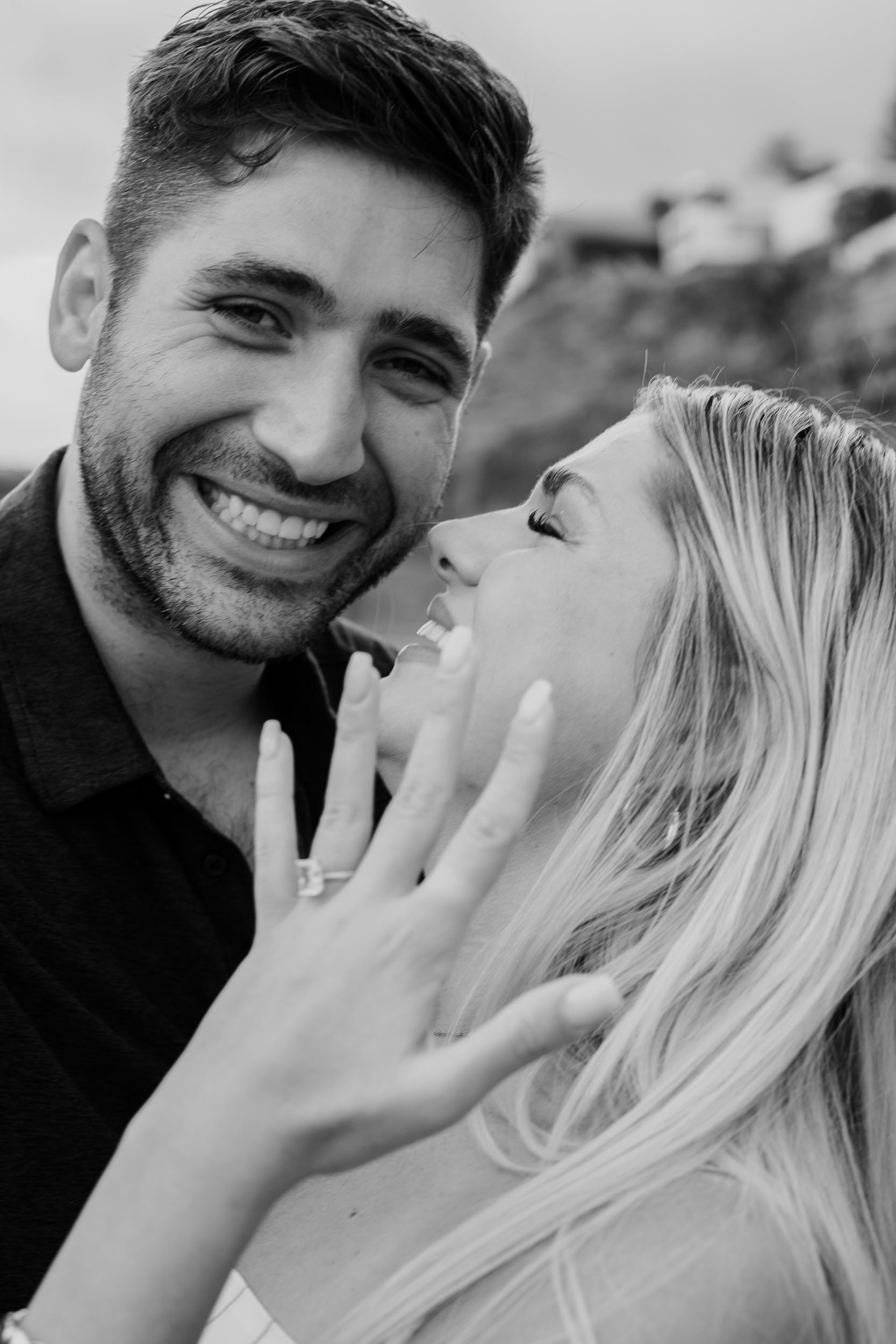 Black-and-white close portrait of Mike grinning straight at the camera while Amelie nuzzles into his cheek with her ringed hand pressed against his jaw, Praia Formosa, Madeira