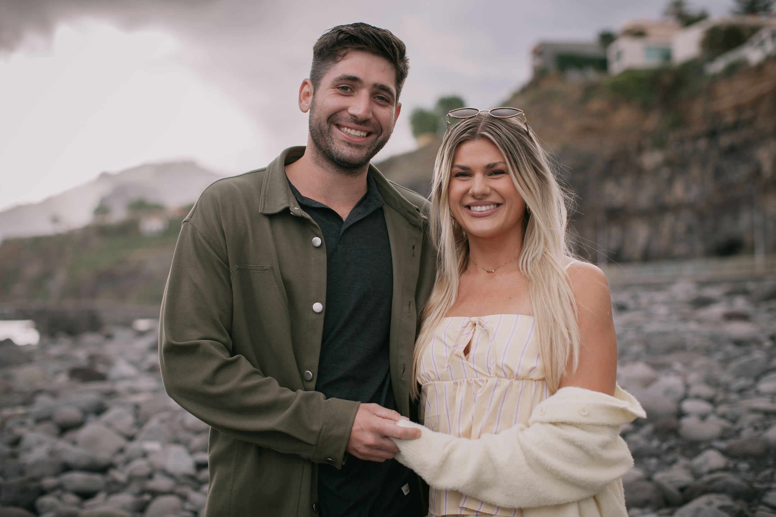 Mike in an olive overshirt and Amelie in a cream cardigan posing together at hand, both smiling straight at the camera, dark volcanic boulders and a cliffside village behind at Praia Formosa, Madeira