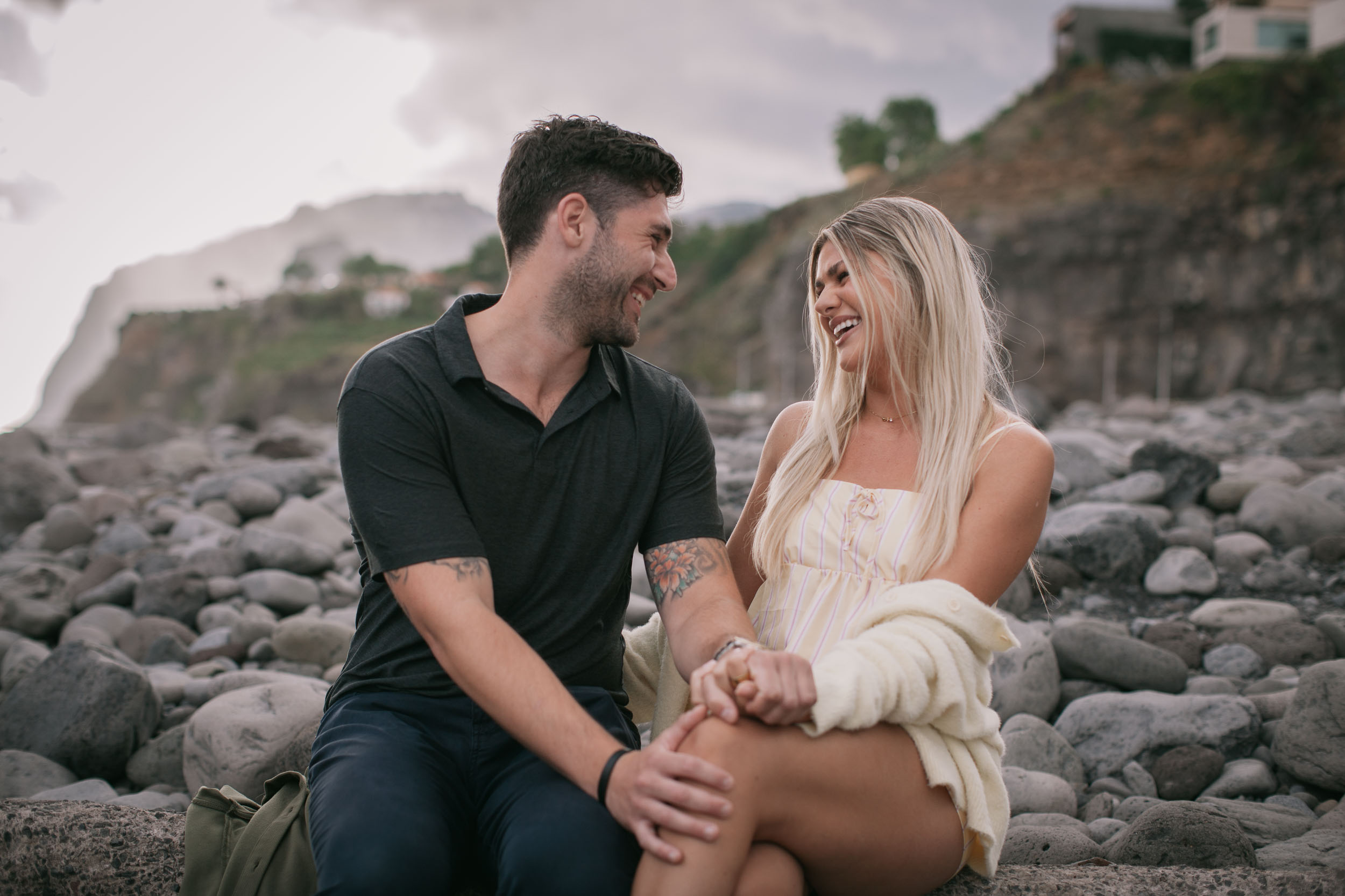Mike and Amelie sitting on a low boulder facing each other and laughing, his hand resting on her bare knee, his small olive backpack on the rocks beside them at Praia Formosa, Madeira