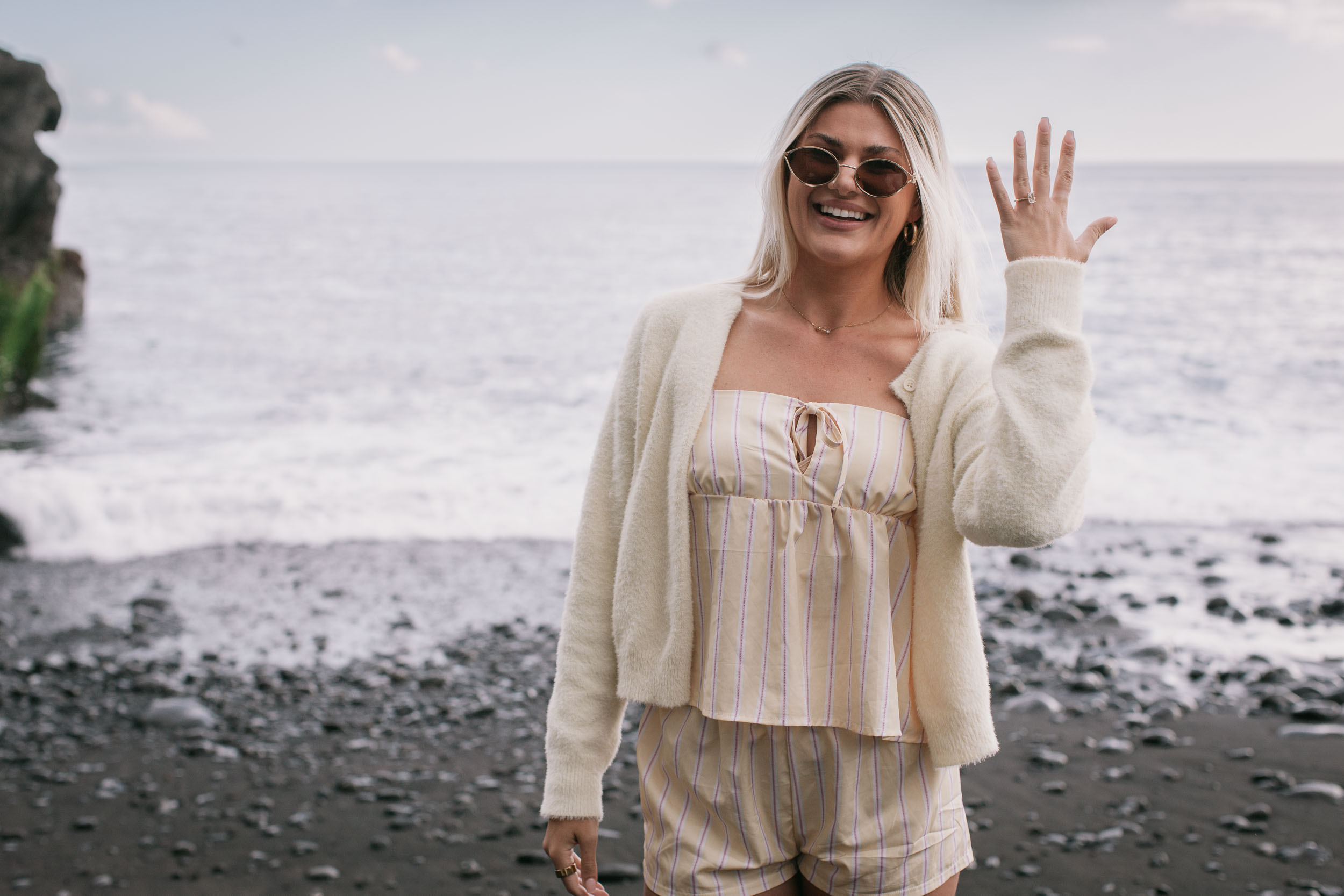 Solo portrait of Amelie standing on the dark sand in round sunglasses and a cream cardigan, raising one hand in a small wave, foaming Atlantic surf and a rock outcrop on the left at Praia Formosa, Madeira