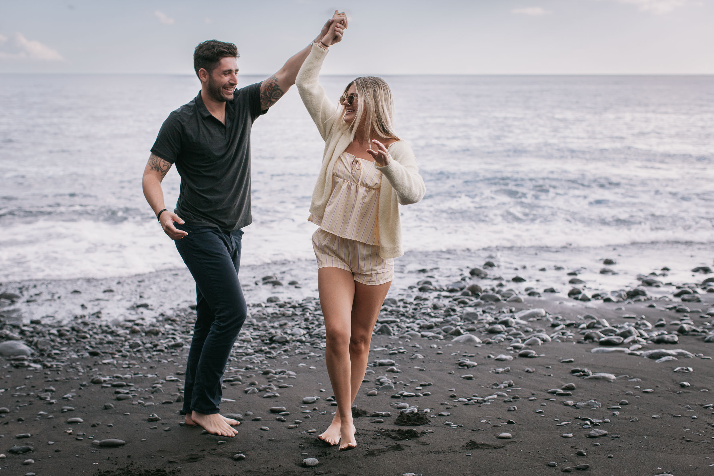 Mike spinning Amelie under his raised arm on the wet black sand, both barefoot and laughing wide, breaking white waves behind them at Praia Formosa, Madeira