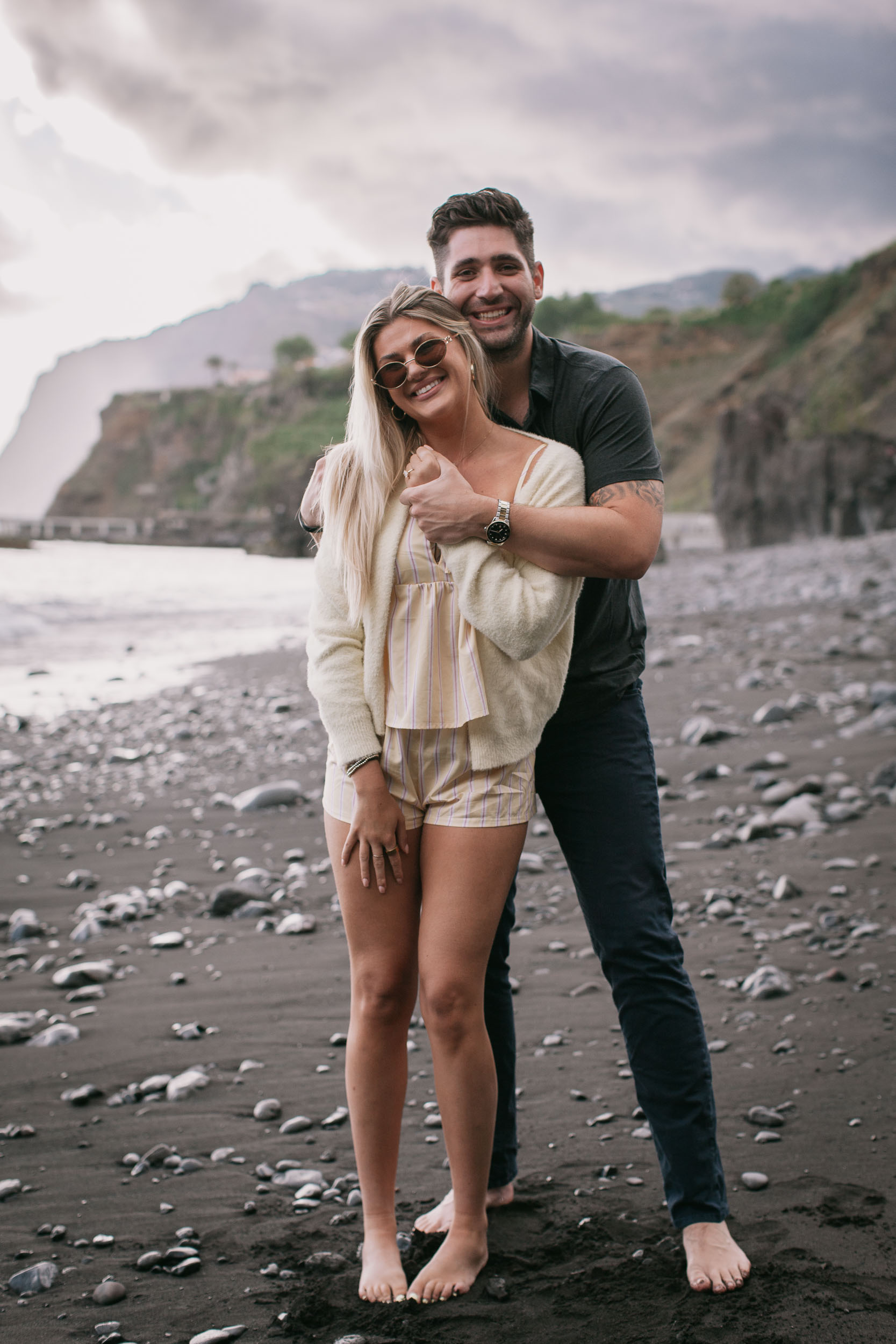 Mike standing behind Amelie with both arms wrapped around her shoulders, both smiling straight at the camera, the green sheer Cabo Girão cliff fading into mist behind them at Praia Formosa, Madeira