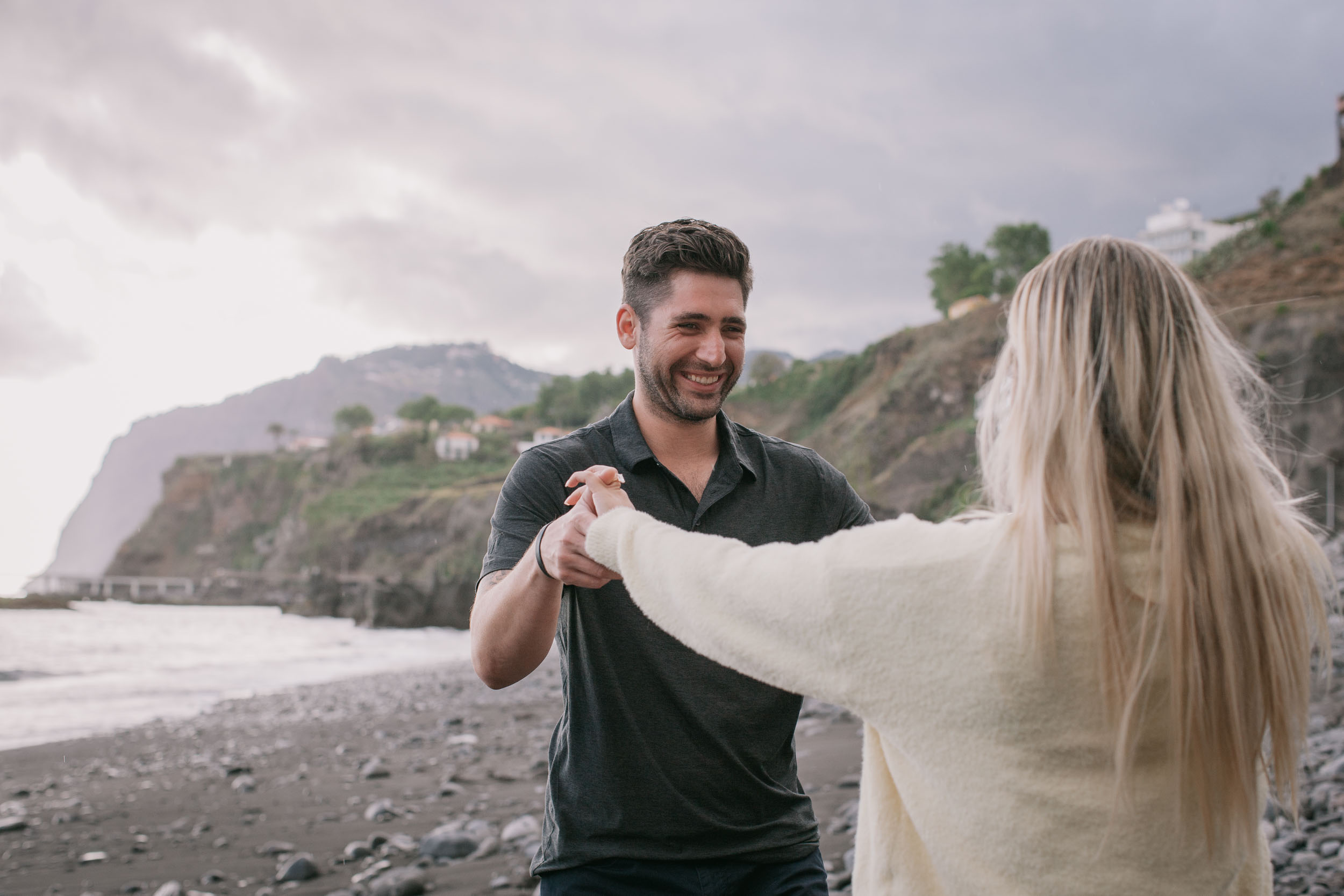Mike laughing as he holds Amelie's hand at arm's length while she stands with her back to the camera, the mountains and white houses of Madeira lit softly behind him at Praia Formosa