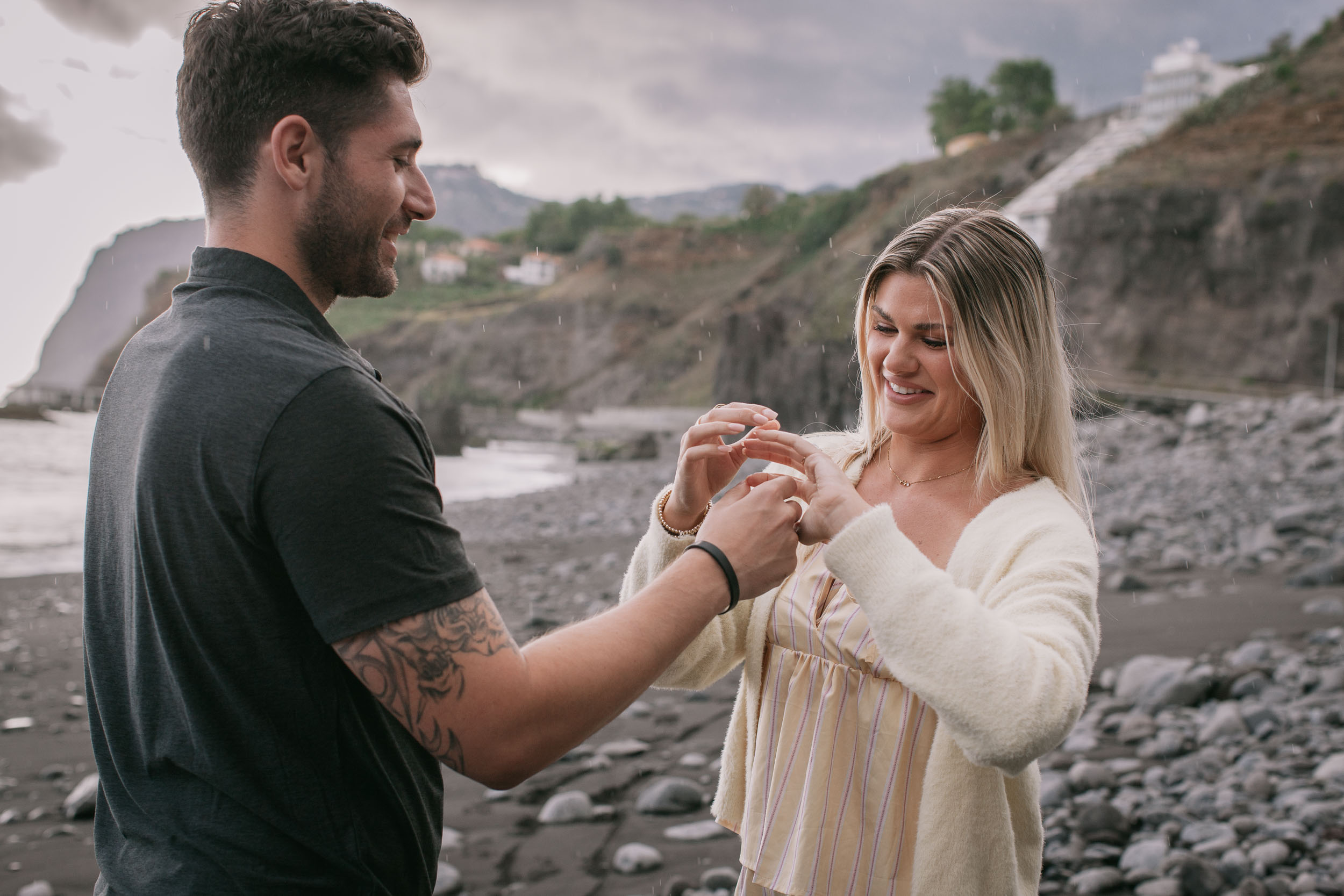 Mike and Amelie standing close as he holds her hand to compare rings between them, light drizzle dotting the air, white cliffside houses behind at Praia Formosa, Madeira