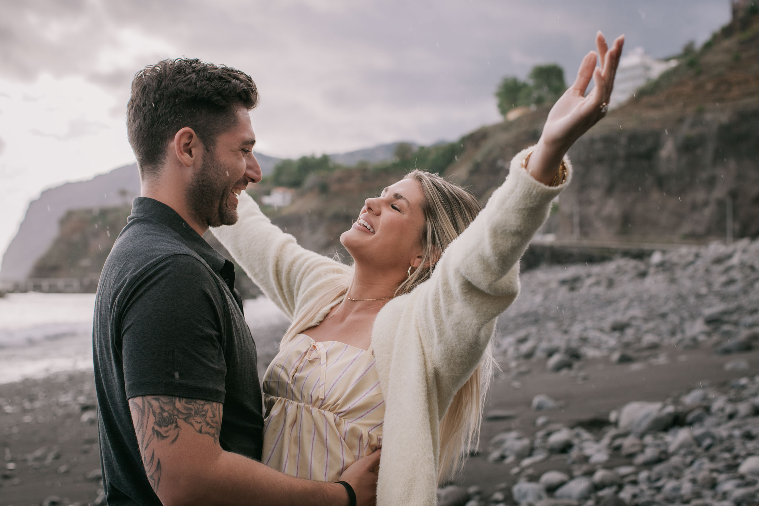 Amelie throwing her free arm up to the sky in joy while Mike laughs and holds her by the waist, light rain streaking the frame, Praia Formosa cliffs softened behind, Madeira