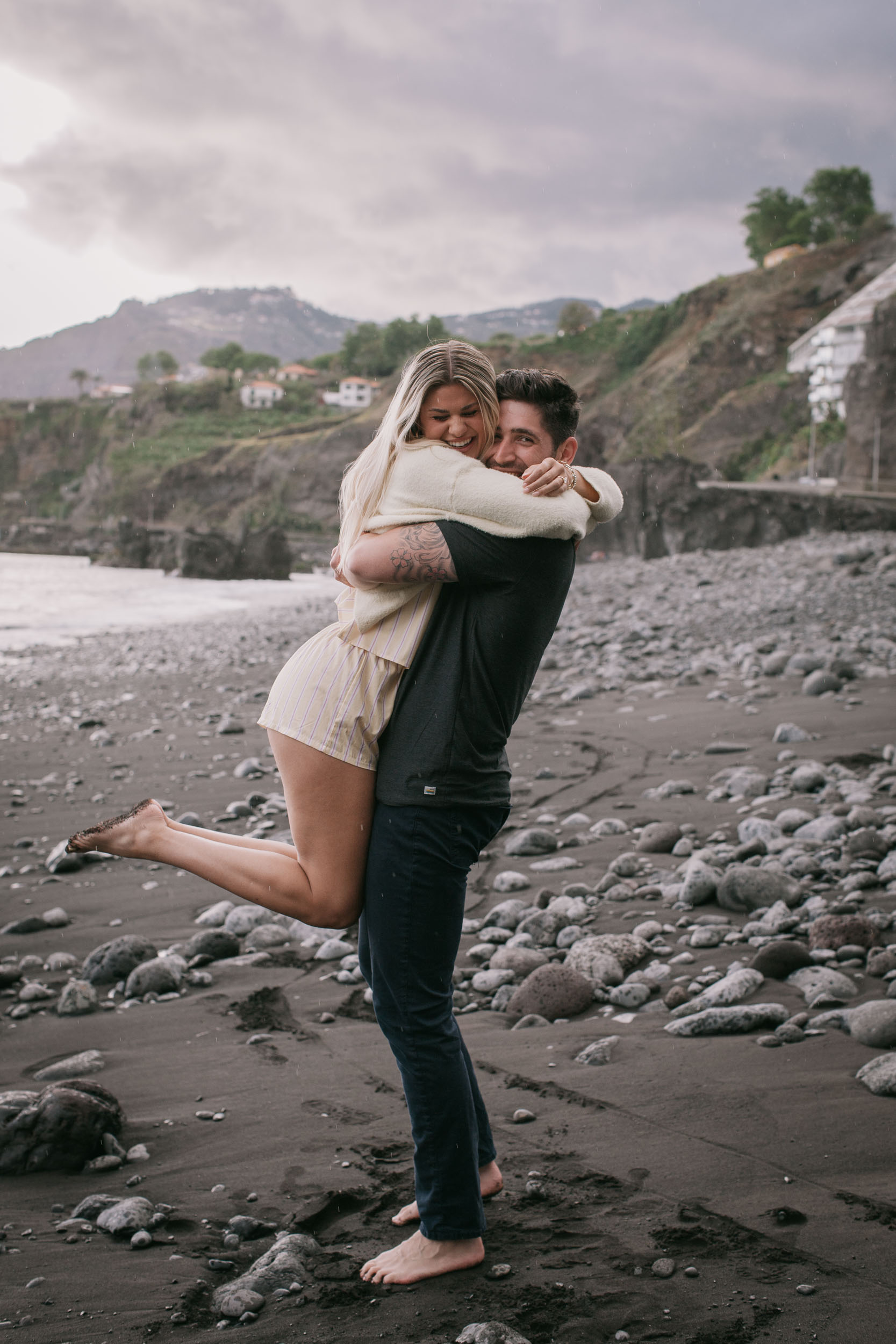 Vertical frame of Mike lifting Amelie off the wet black sand in a tight hug, her bare leg kicked high behind her, the mountain village and terraced slopes filling the background at Praia Formosa, Madeira