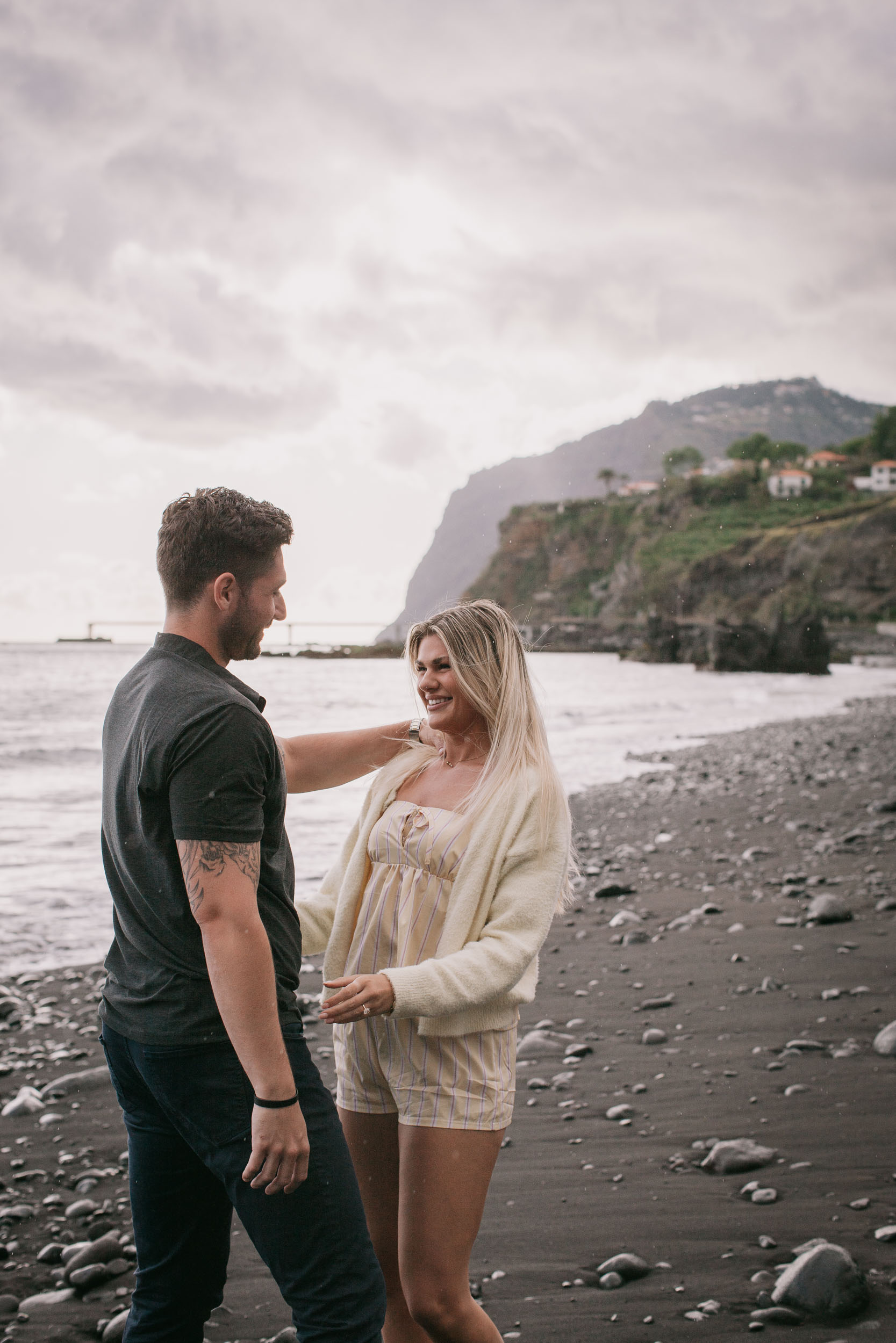Mike resting a hand on Amelie's shoulder as she looks up at him smiling, both standing on the wet sand at the shoreline, the steep Funchal cliff and red-roofed village behind at Praia Formosa, Madeira