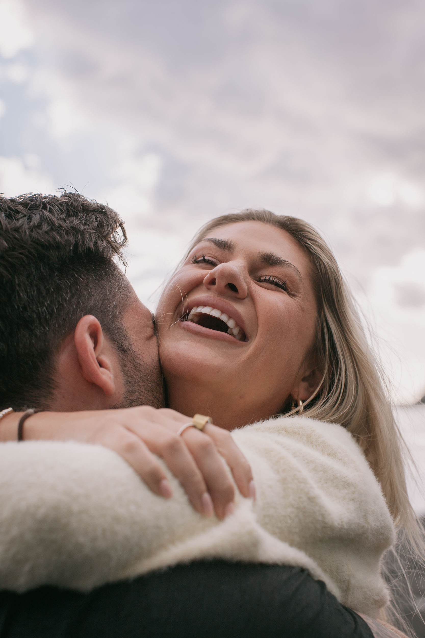 Low-angle vertical of Mike kissing Amelie's cheek from behind, her face tipped up with eyes closed in a wide laugh, drizzle and lavender clouds filling the sky at Praia Formosa, Madeira