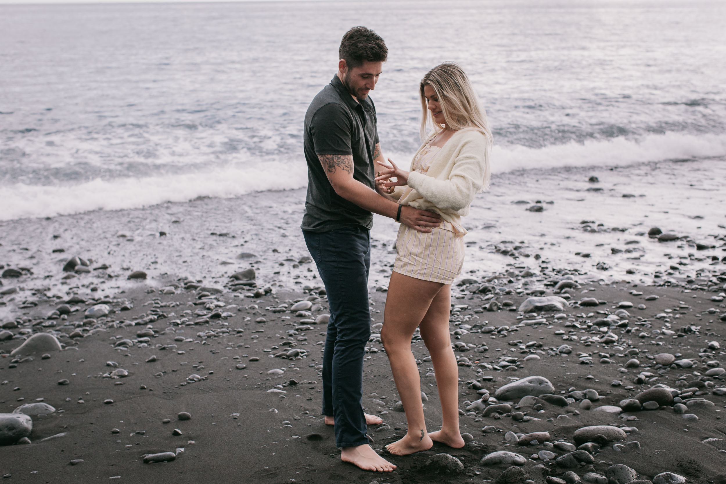 Mike and Amelie standing barefoot at the water's edge both looking down at where his hands hold her hand, foaming surf curling beside them at Praia Formosa, Madeira