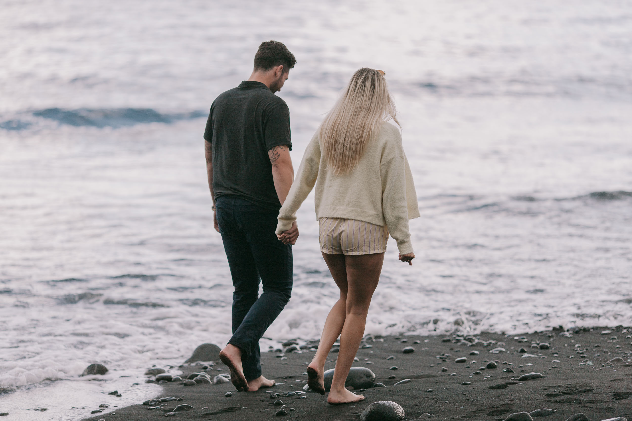 Mike and Amelie seen from behind walking hand in hand into the shore break, his dark t-shirt and her cream sweater bright against the moving Atlantic at Praia Formosa, Madeira