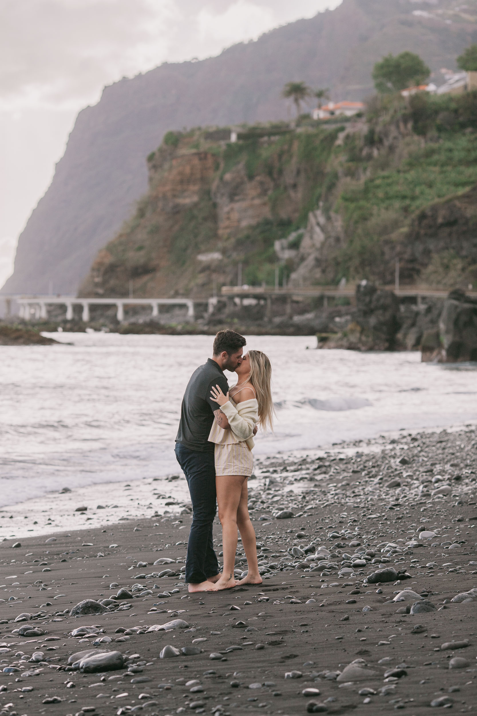 Vertical of Mike and Amelie in a long quiet kiss on the wet black sand, the green sheer face of Cabo Girão and the coast road viaduct filling the entire background at Praia Formosa, Madeira
