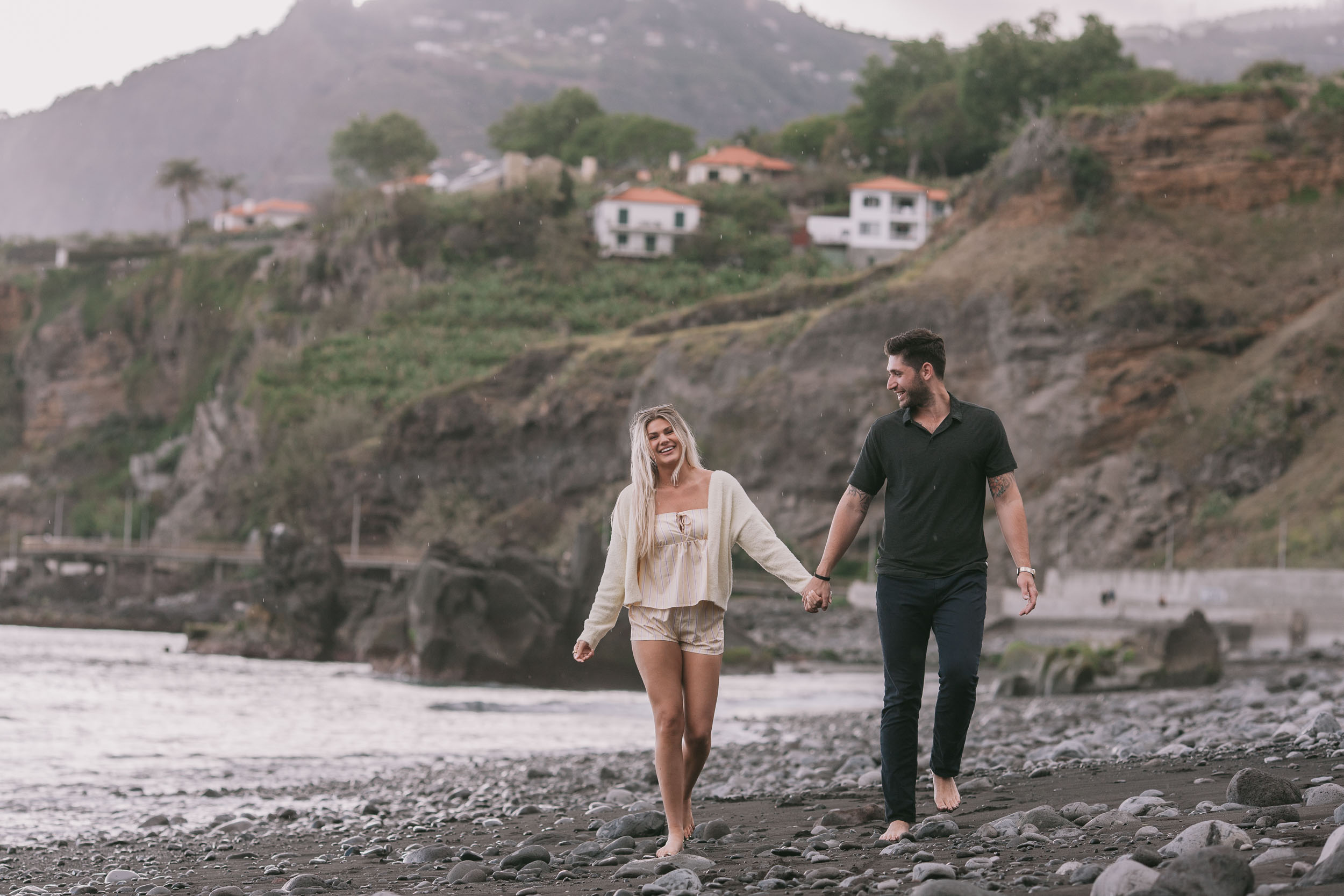 Amelie smiling back at Mike as they walk hand in hand toward the camera along the boulder shore, the bright white village of Câmara de Lobos climbing the cliff behind them at Praia Formosa, Madeira