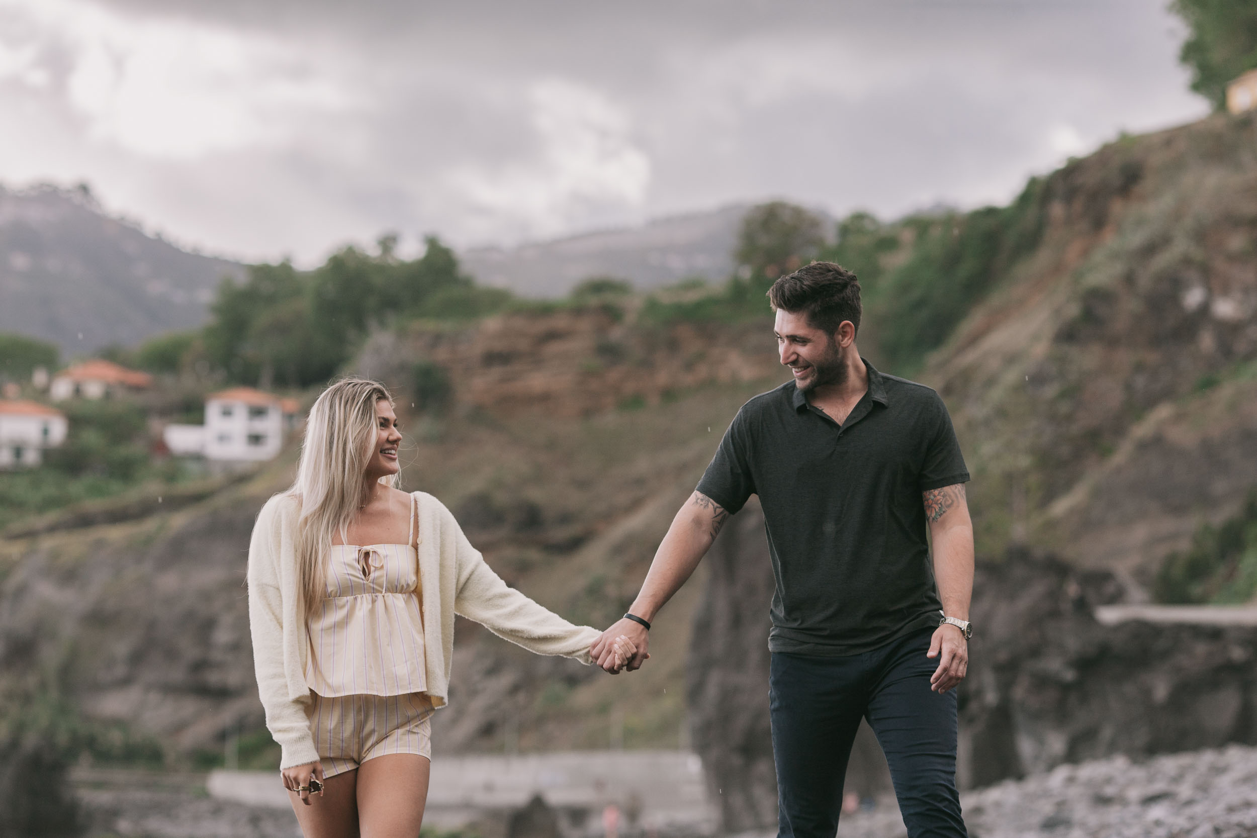 Mike and Amelie walking toward the camera with fingers barely linked, his black polo and her cream cardigan against the brown terraced hillside and a white house far behind at Praia Formosa, Madeira