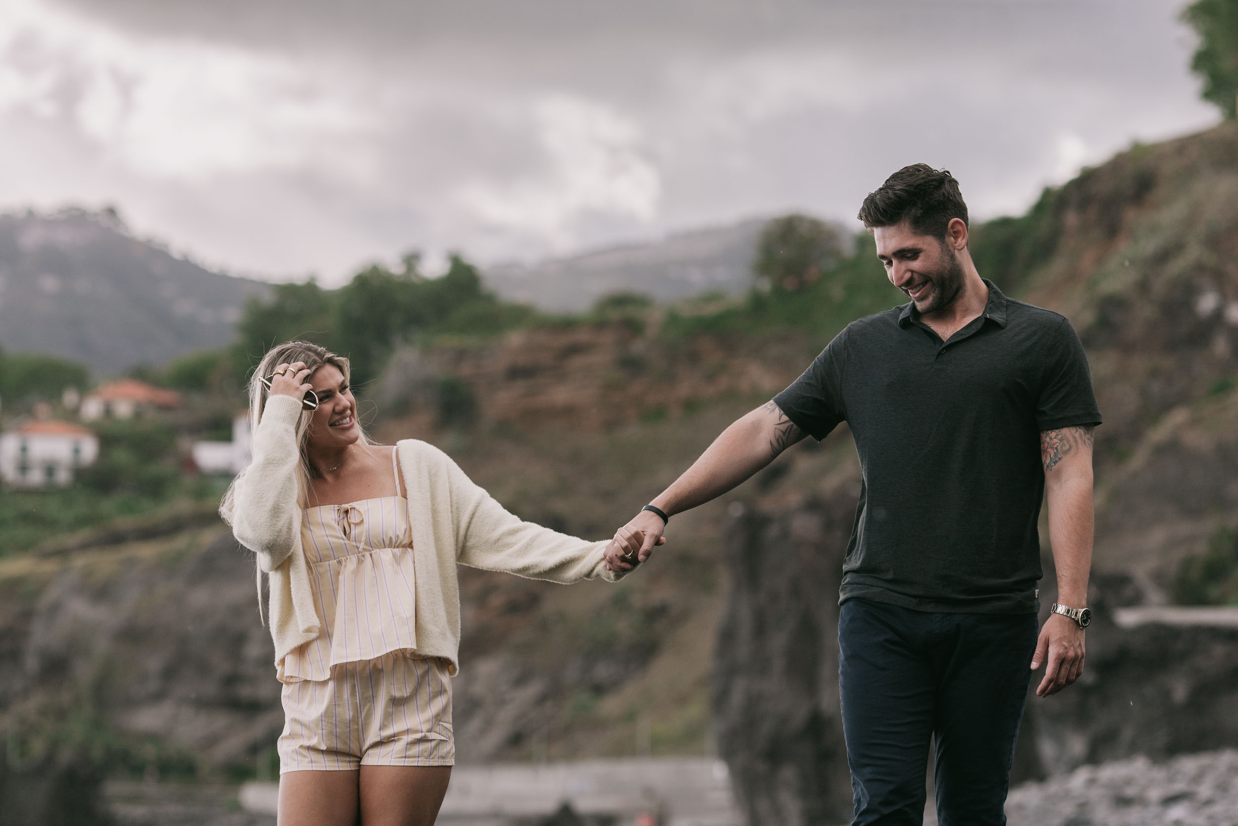 Amelie shielding her eyes with one hand while holding Mike's hand and walking beside him, both smiling toward each other, the white house and terraced slope behind at Praia Formosa, Madeira