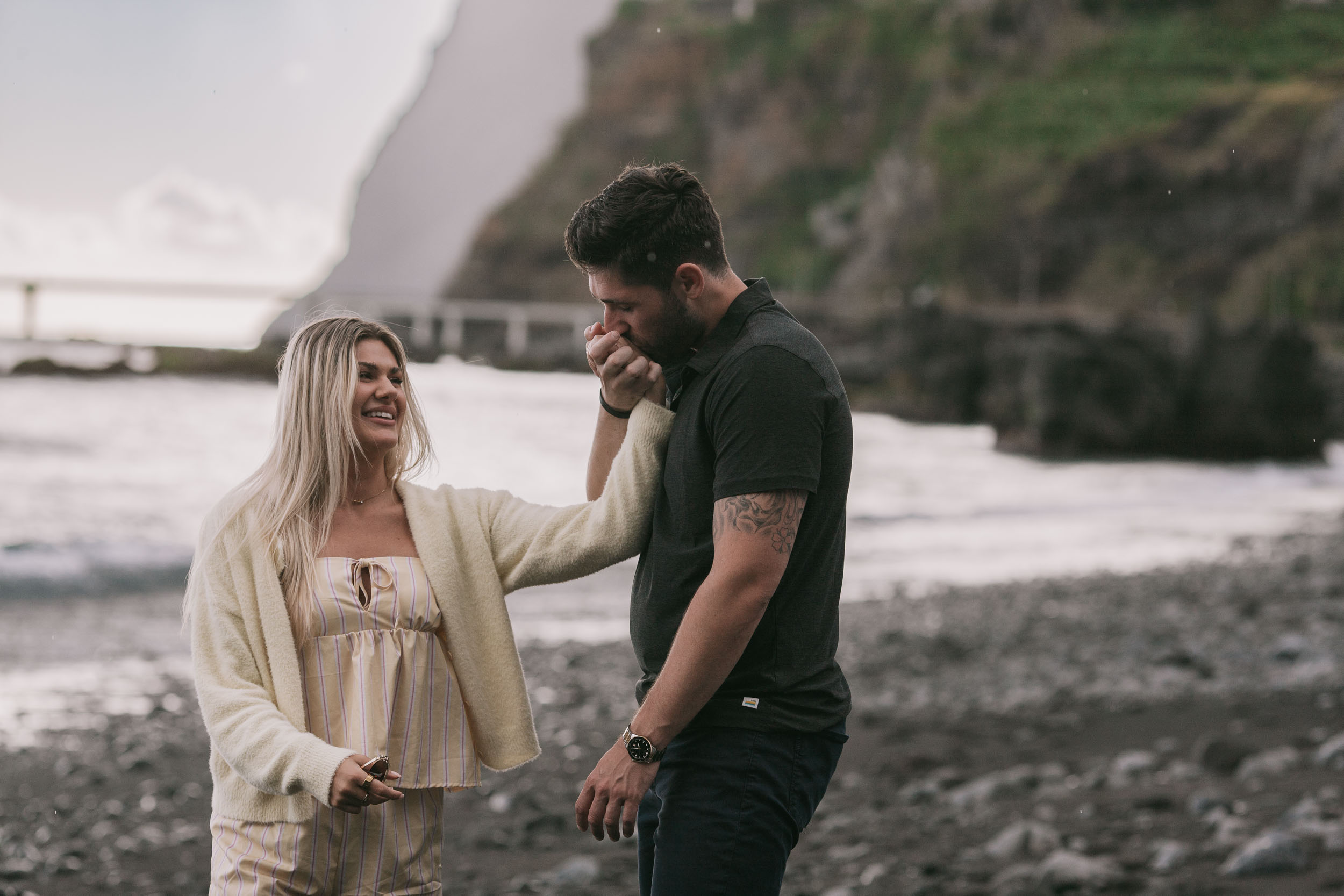 Mike bowing slightly to kiss the back of Amelie's hand while she laughs and holds the small dark ring box in her other hand, ocean spray and the Funchal bridge softened behind at Praia Formosa, Madeira