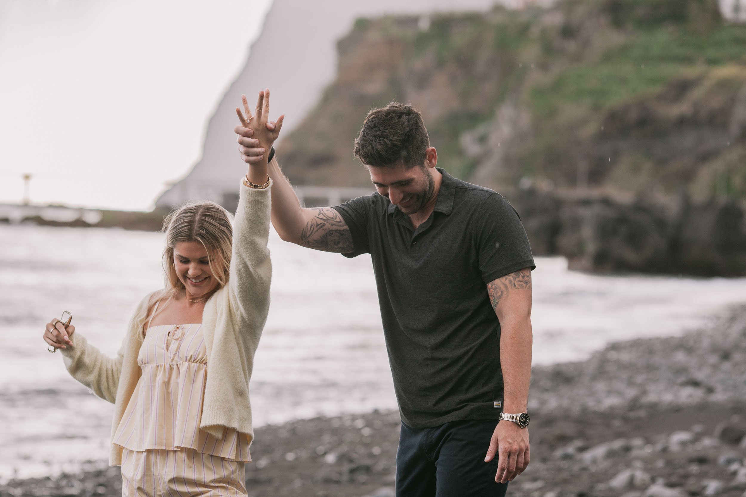 Amelie laughing wide as she ducks under Mike's raised arm in a twirl, her bracelets sliding down her arm and his watch on his wrist, light rain in the air at Praia Formosa, Madeira