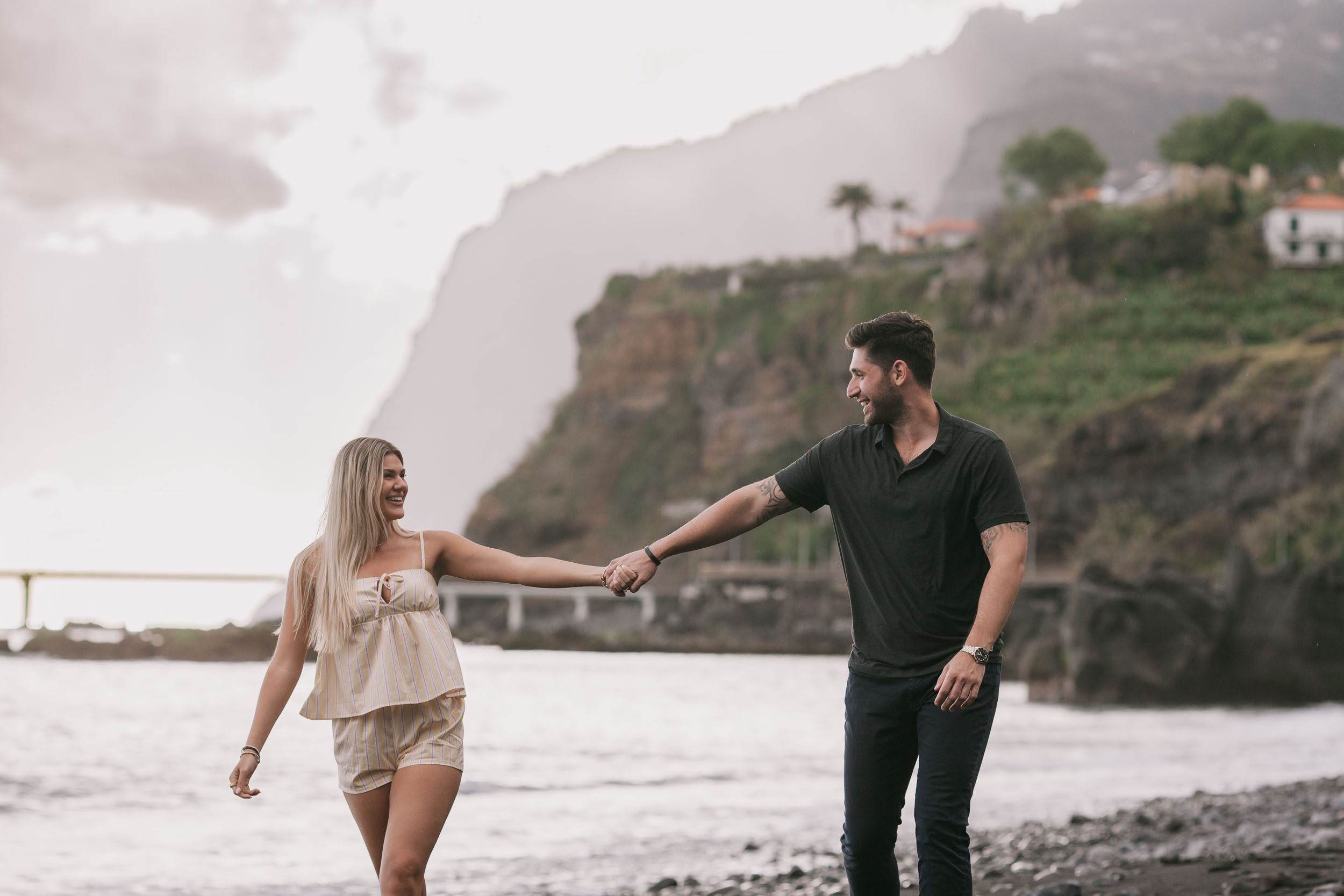 Mike grinning back at Amelie as she walks behind him pulling on his hand, the sheer green wall of Cabo Girão filling the background at Praia Formosa, Madeira