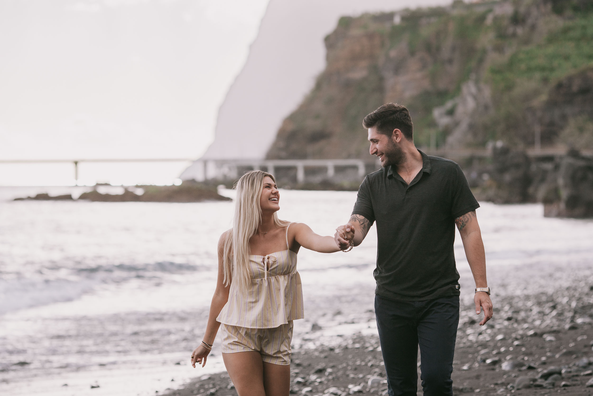 Mike and Amelie walking side by side hand in hand on the wet pebble shore, her hair lifting in the breeze, the dim profile of Cabo Girão and the bridge behind at Praia Formosa, Madeira