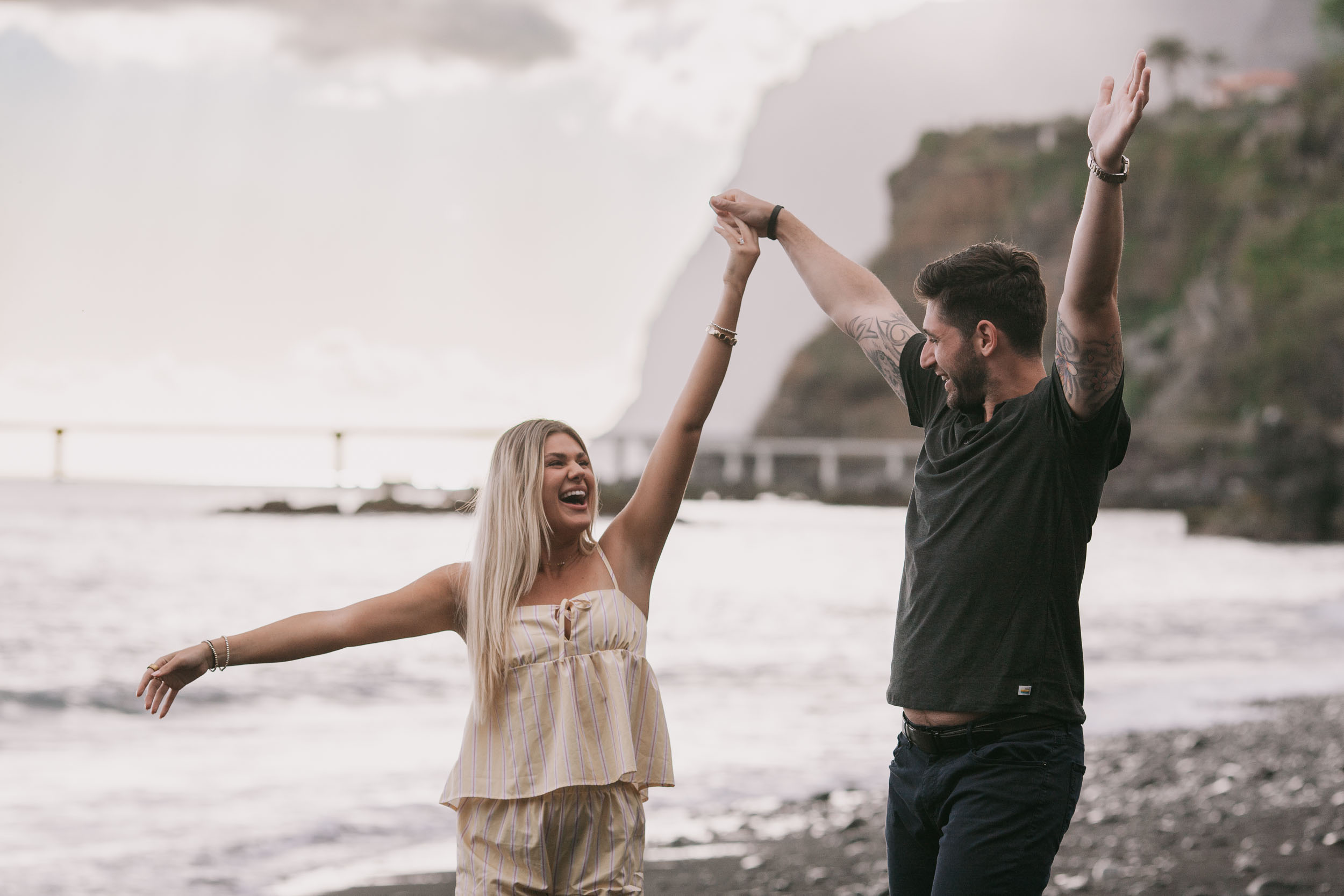 Mike and Amelie both throwing their arms up over their heads in a single victorious gesture, hands meeting at the top, the green Cabo Girão cliff in soft mist behind at Praia Formosa, Madeira
