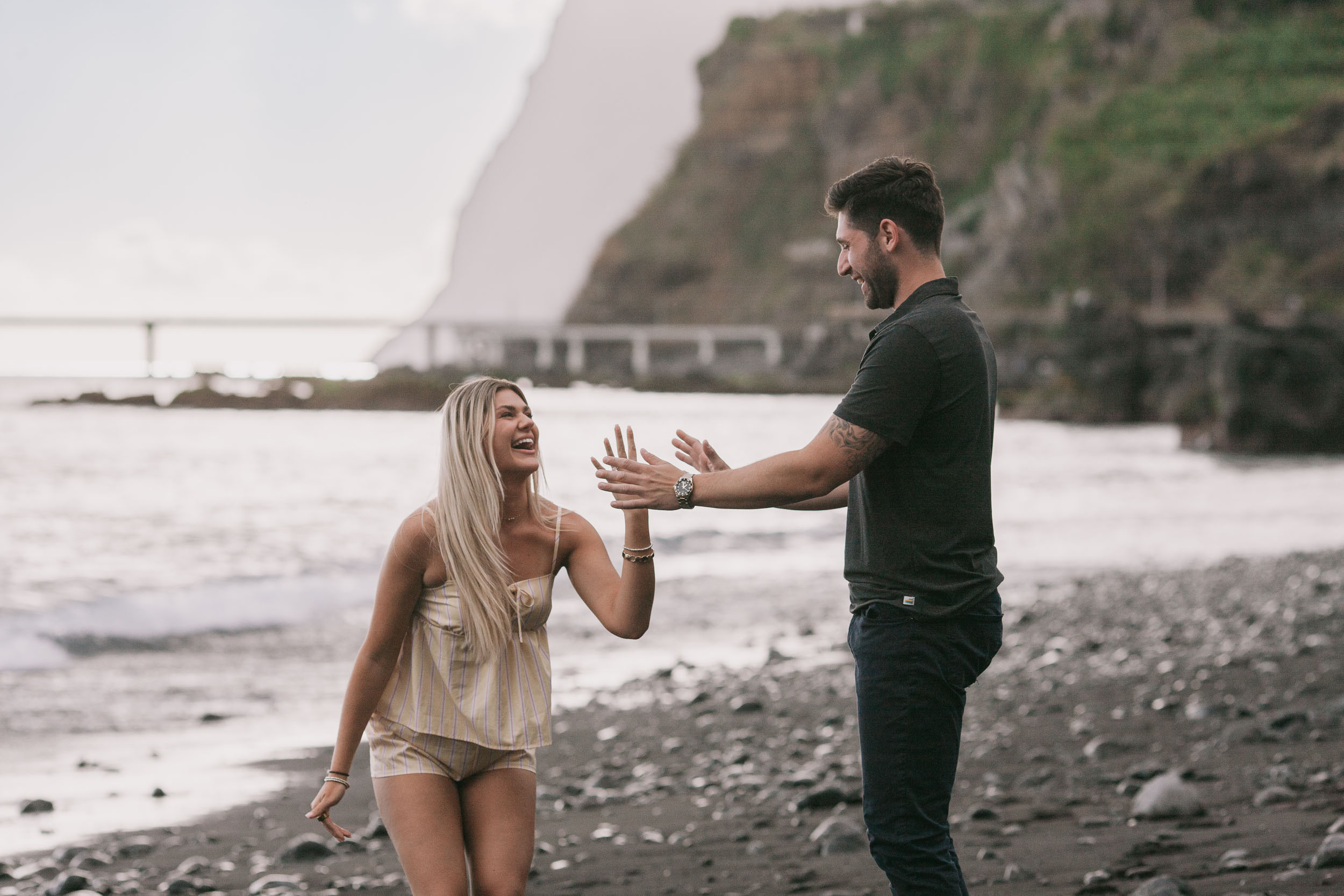 Amelie laughing and stretching both hands forward toward Mike as he reaches back toward her on the wet sand, the cliff and bridge softened behind at Praia Formosa, Madeira
