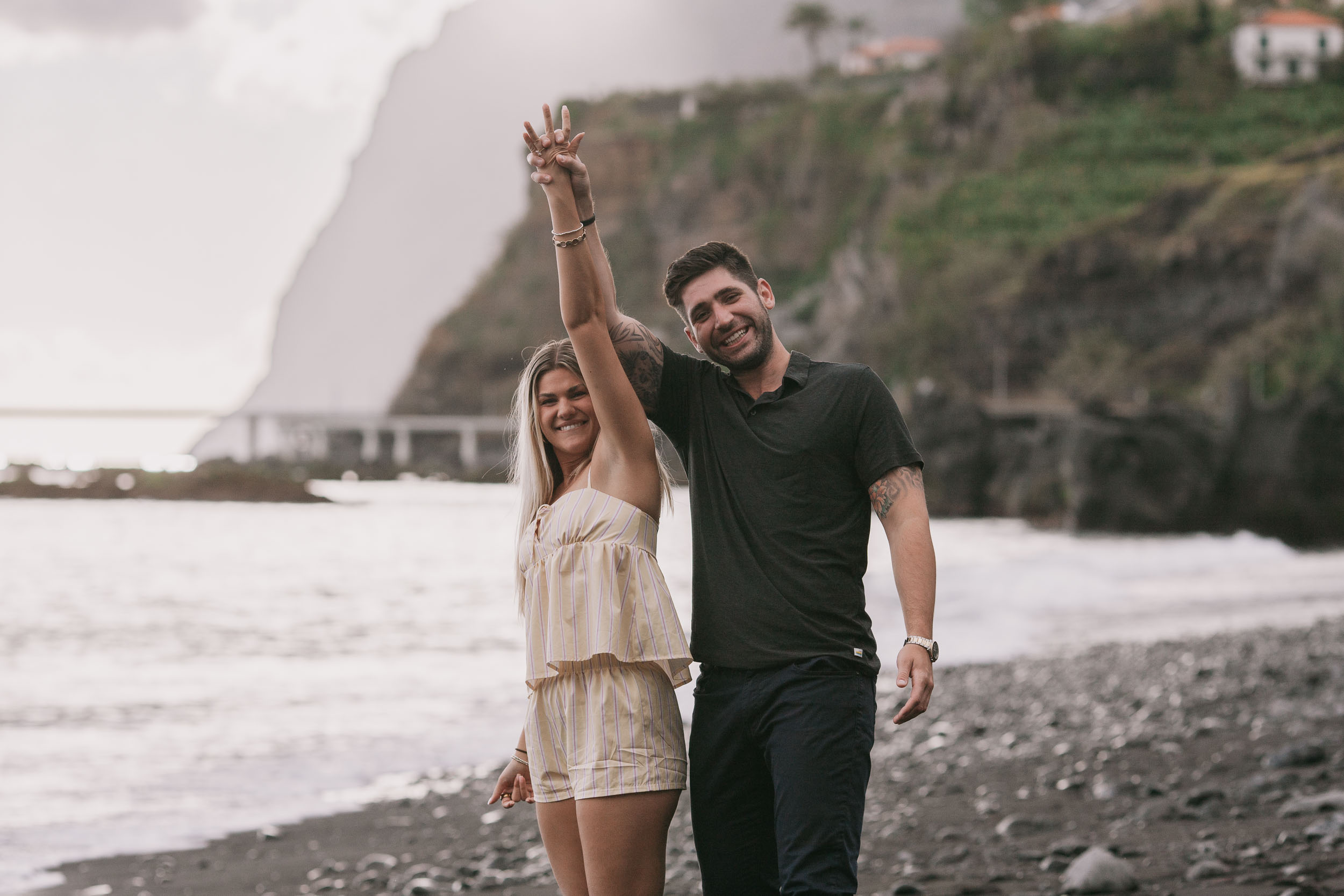 Amelie throwing up a single peace sign while Mike grins beside her with one arm around her shoulder, both looking straight at the camera, the misty Cabo Girão cliff behind at Praia Formosa, Madeira